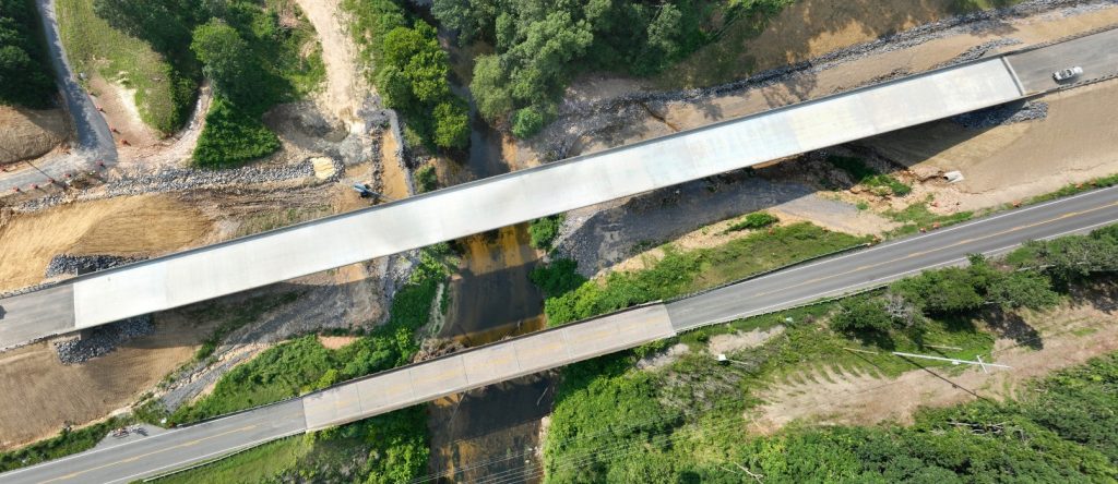 An overhead view of two bridges crossing a creek shows the much greater size of the new bridge, top, compared to the old bridge, bottom.