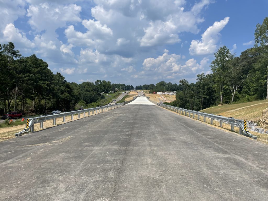 A new roadway, lined by shiny new guardrail, approaching a new concrete bridge surface.
