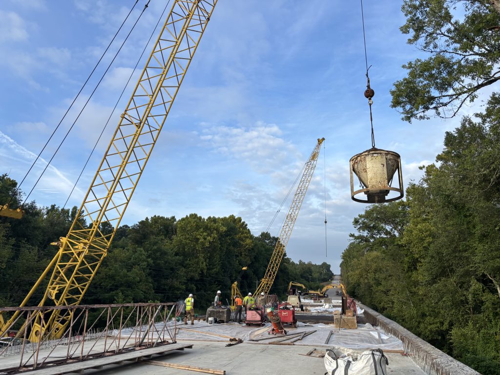 A crane hoists a large metal container above a bridge. A crew of workers are standing on the bridge. Another crane is visible in the background.