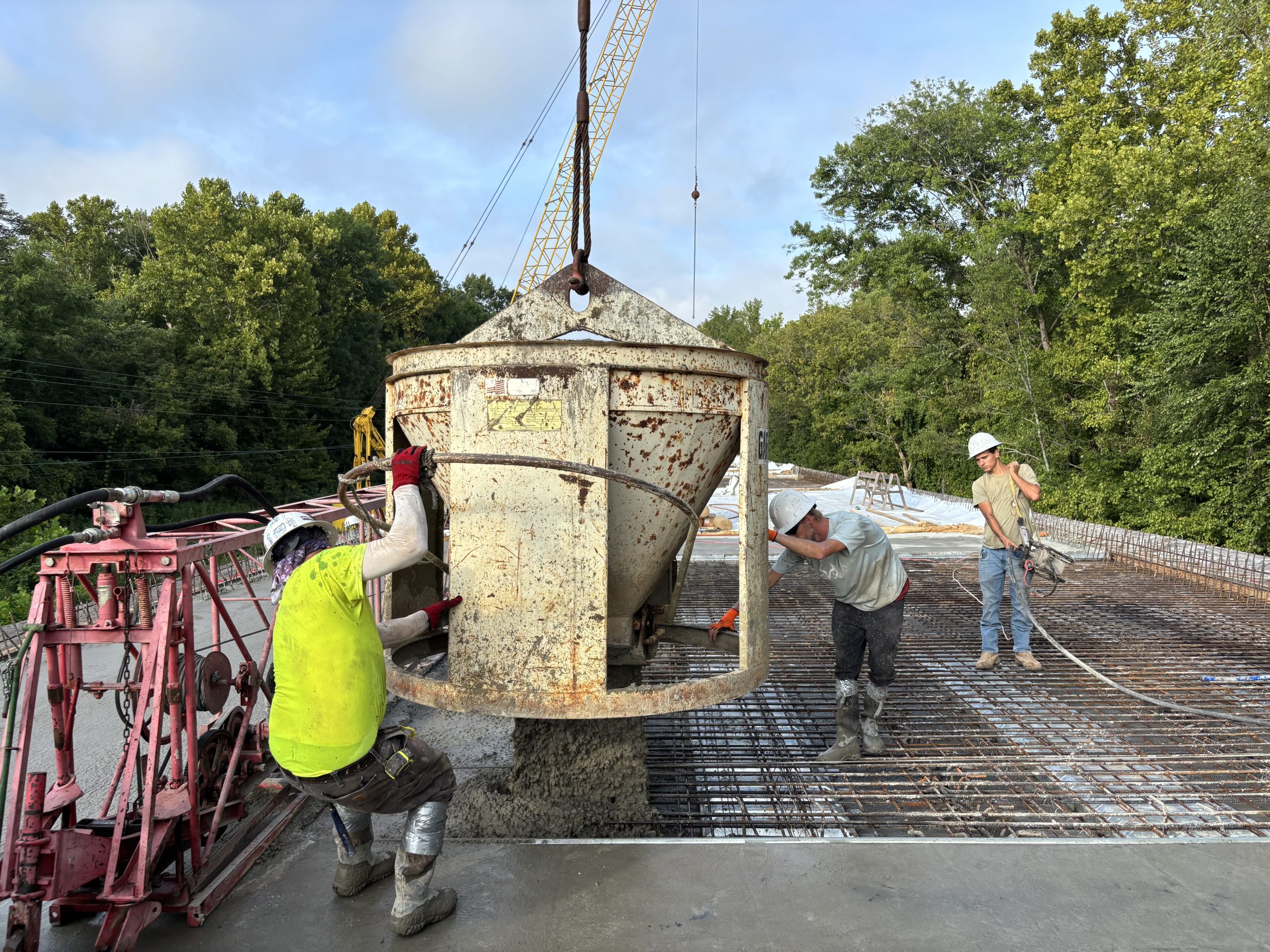 Workers in hard hats manuever a large metal bucket suspended by a crane to pour concrete over rebar.