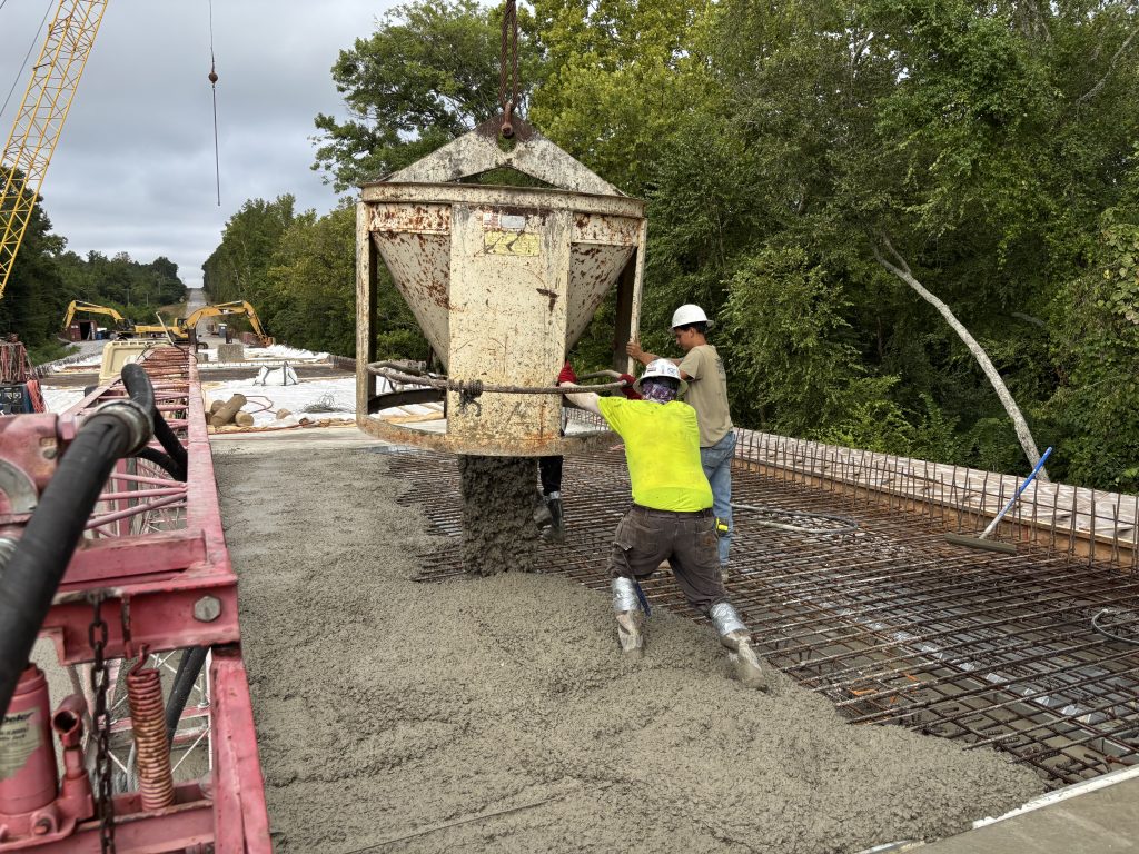 Workers direct a large metal bucket from which wet concrete pours.