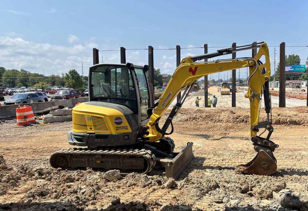 An excavator with a row of pilings and a group of men in yellow vests having a discussion visible in background