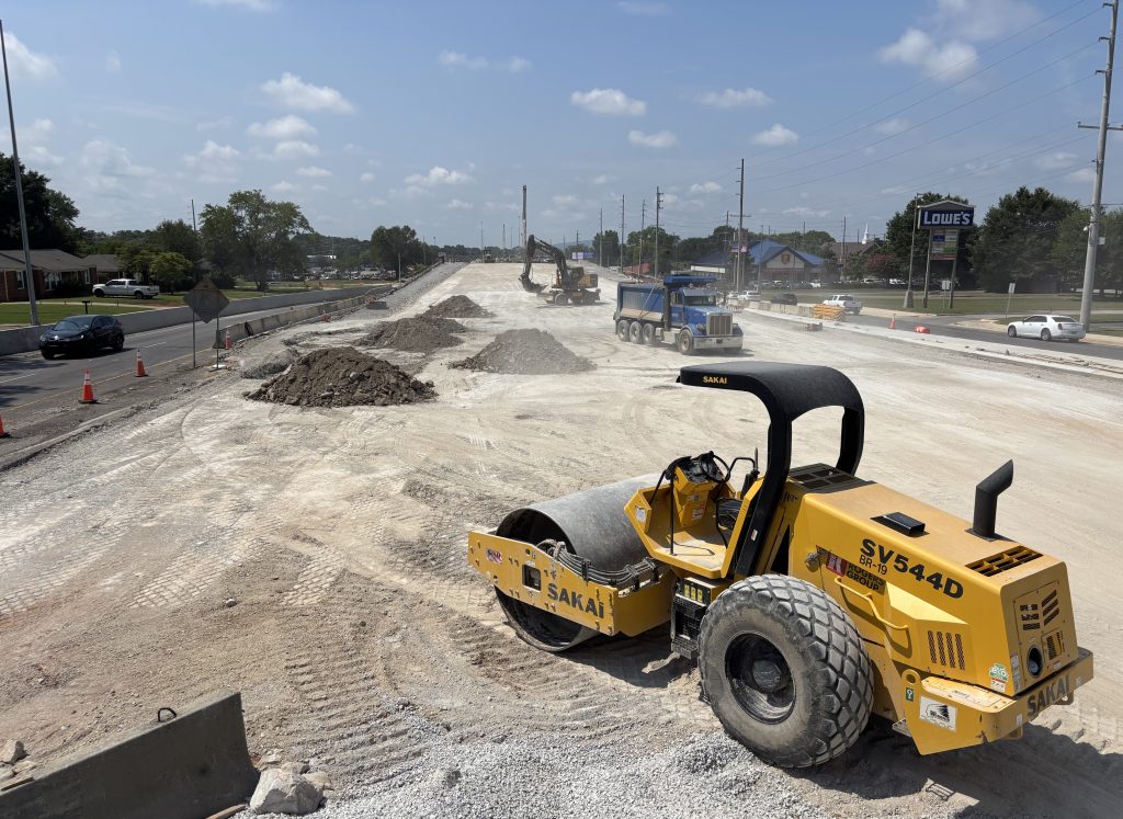 A construction site with a road roller in the foreground and a dump truck and an excavator visible in the background.