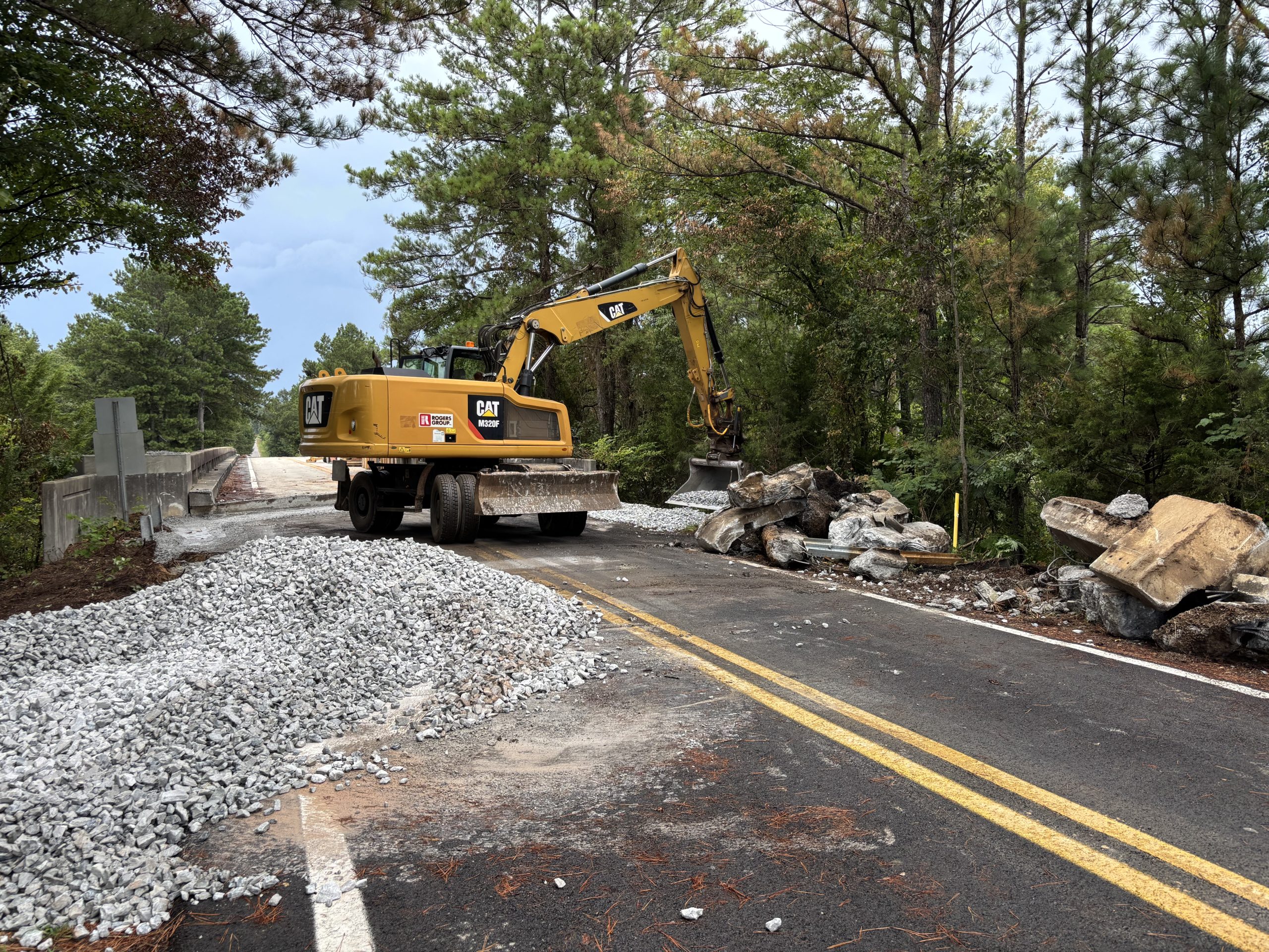 A pile of stone sits in the foreground. An excavator pours stone from its shovel onto the shoulder of a road. The surface of the bridge in the background is higher than the road surface.