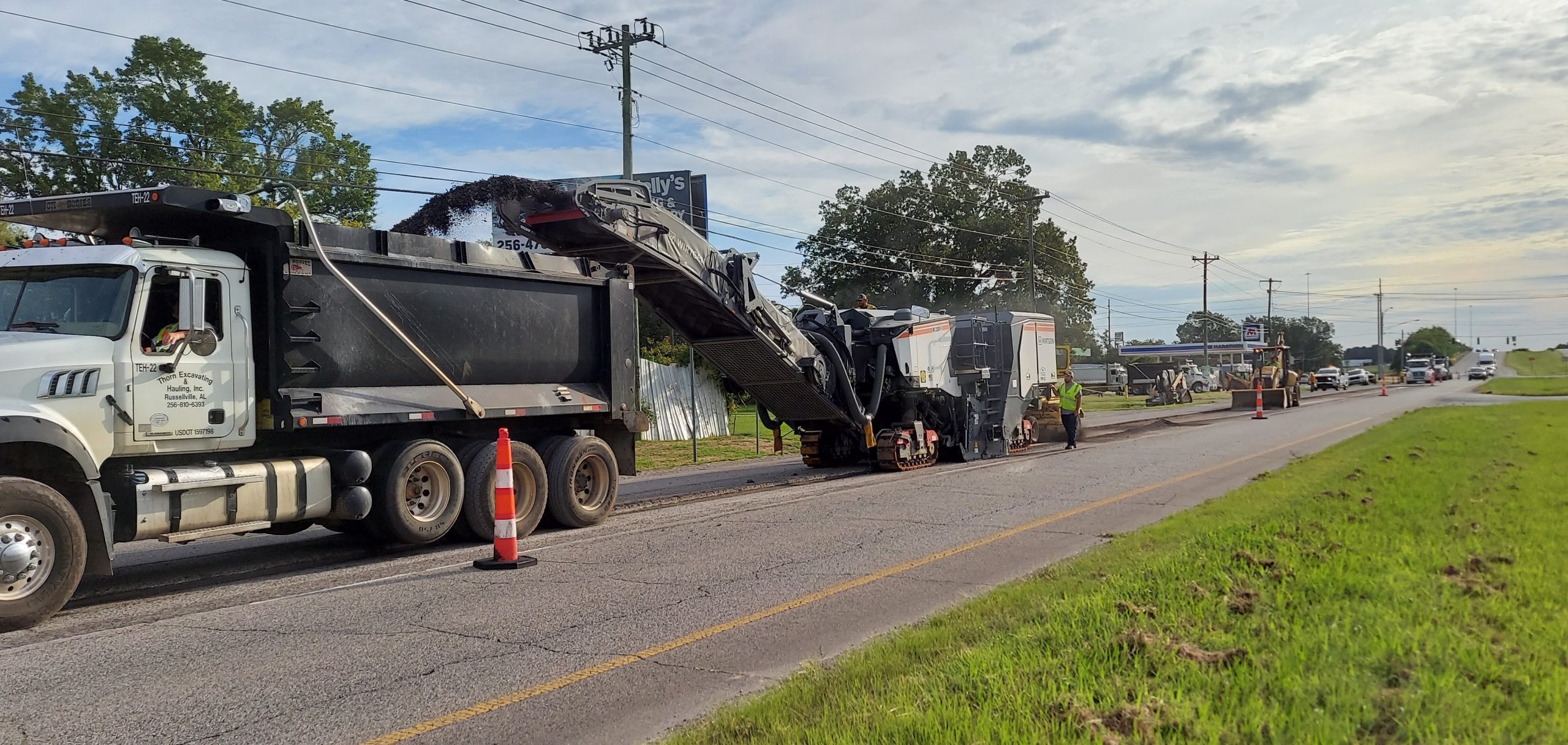 A milling machine conveys asphalt millings into a dump truck on the outside lane of a highway. A sweeper and a front-end loader follow behind them to remove remaining dust and debris.