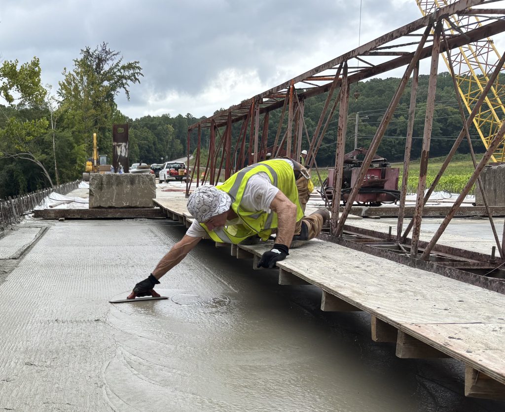 A worker leans from a platform to smooth concrete that has been leveled by a screed.