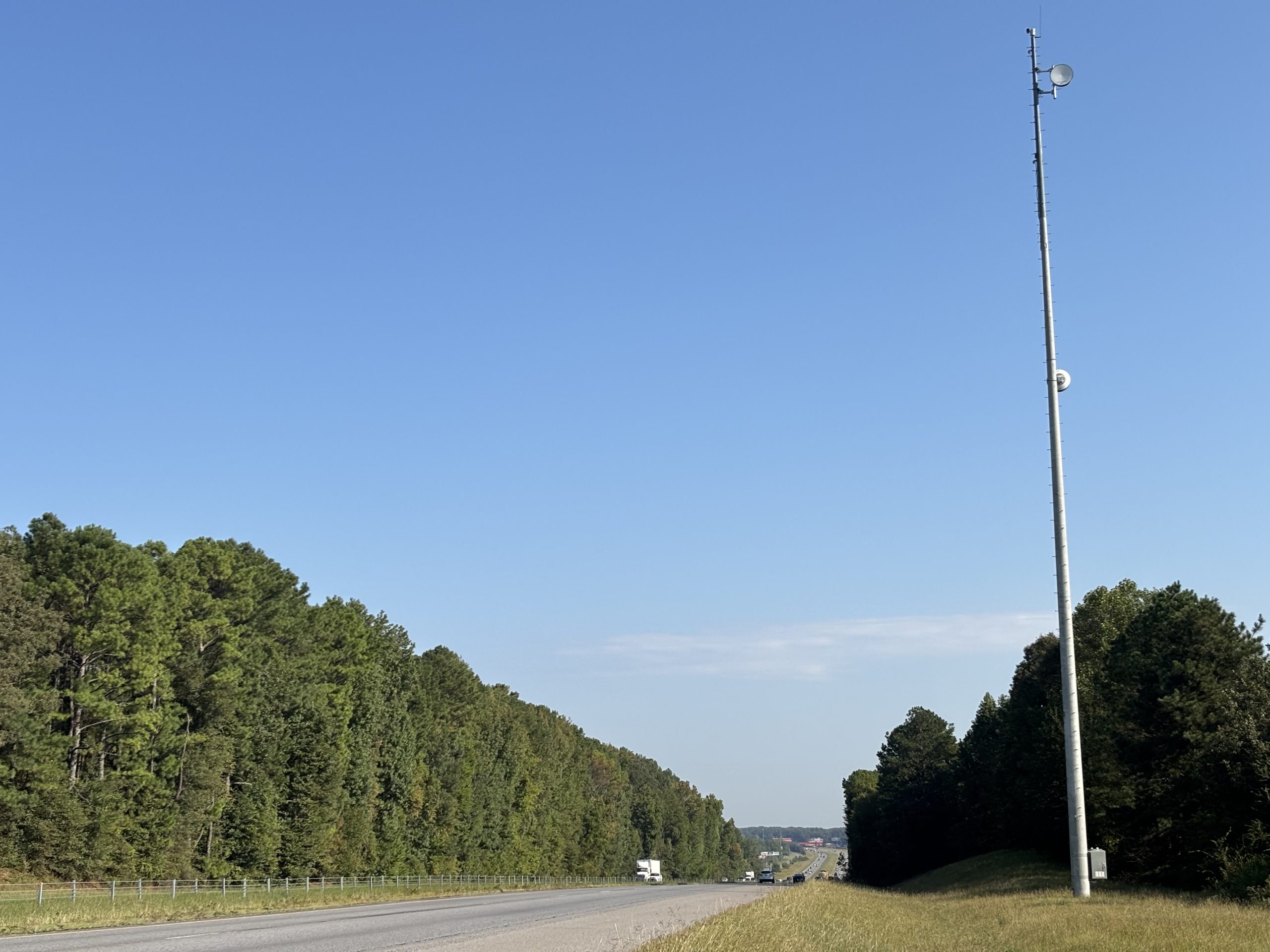 A camera mounted on a tall pole stands on the right of way near an interstate highway. A commercial truck is traveling on the road in the distance.