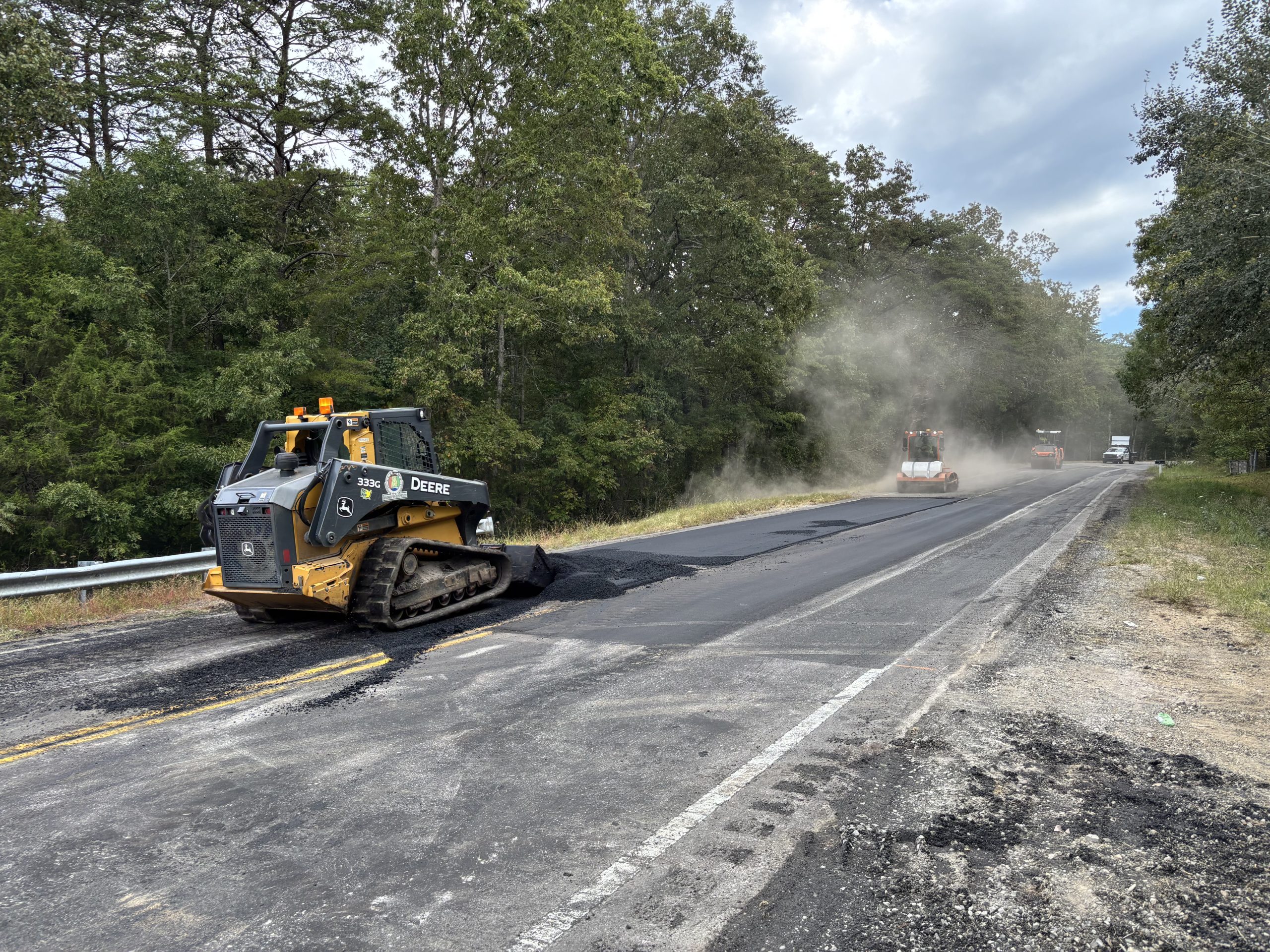 A skid steer and a sweeper operating on a two-lane highway, which is partly freshly paved.