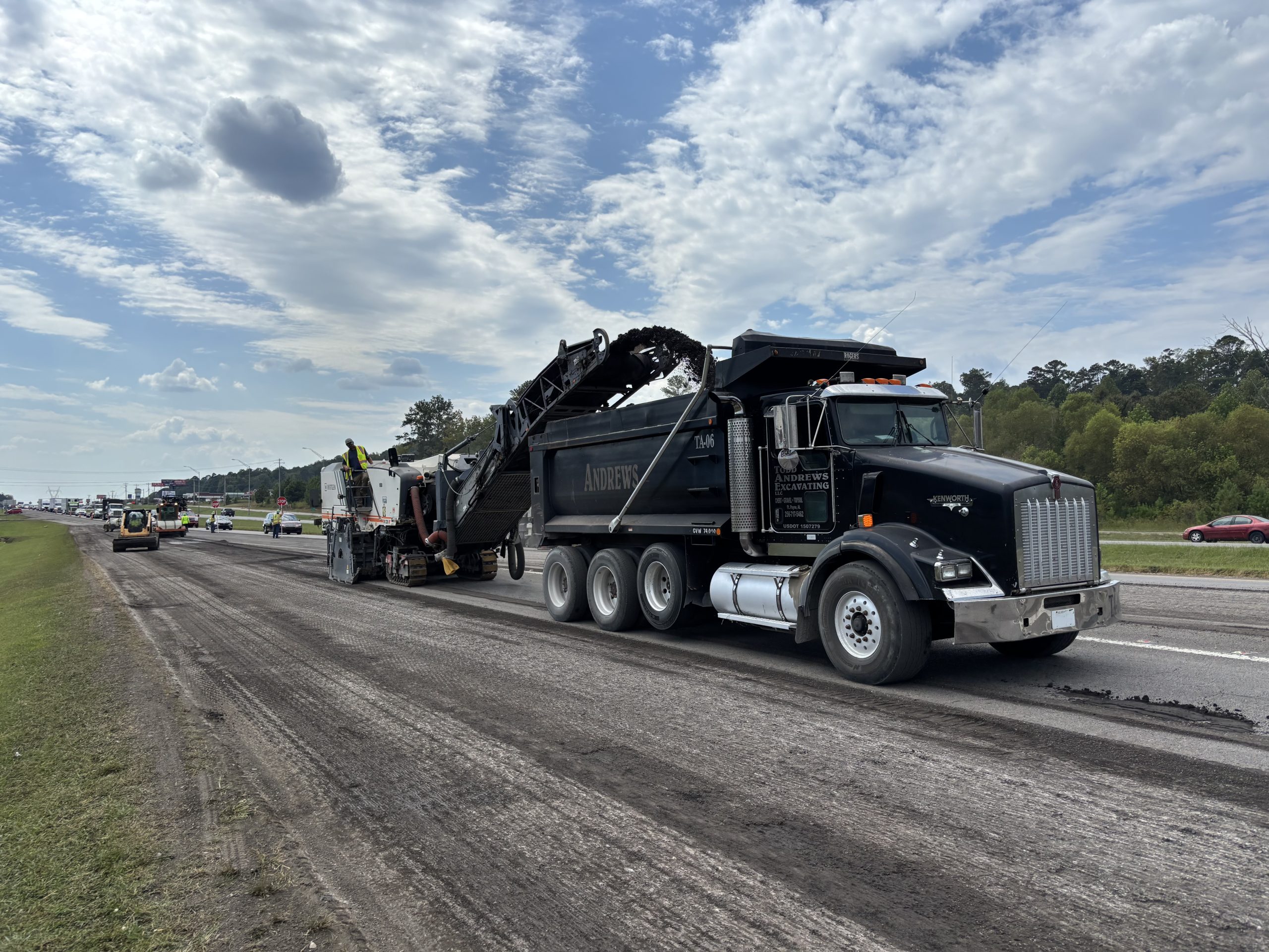 A milling machine deposits millings in a dump truck. Other machinery can be seen working on the milled highway in the background.