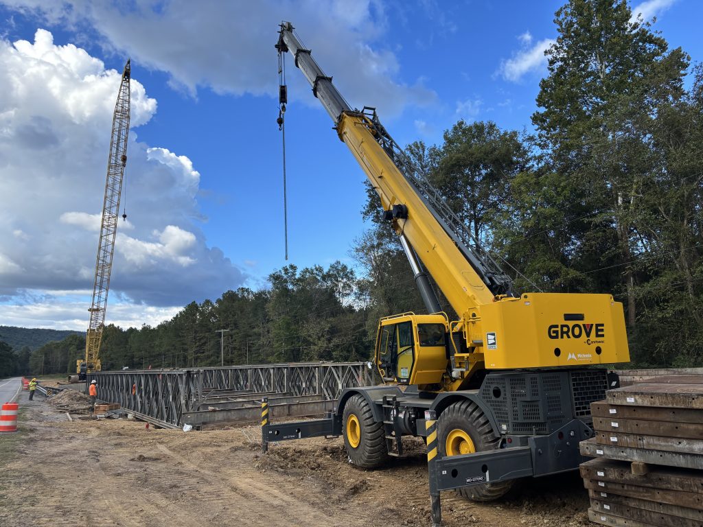 With a crane truck in the foreground and a larger crane in the background, workers assemble a modular bridge in between.