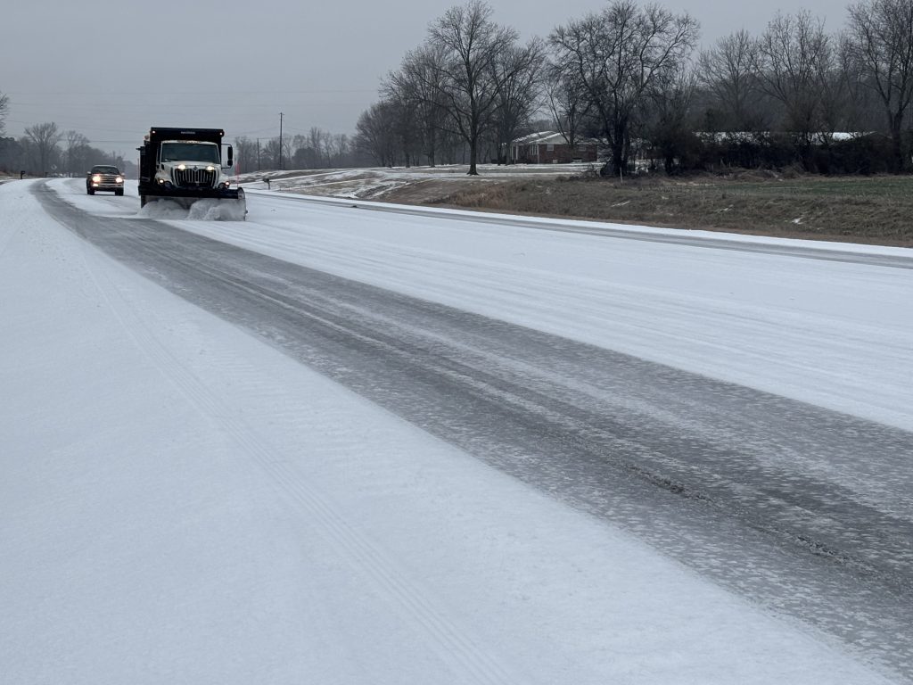 A dump truck with a snow plow mounted on the front scrapes sleet from a roadway.