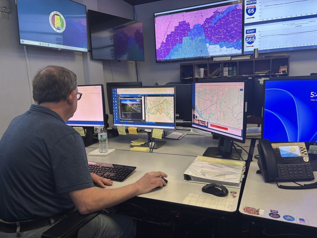 A TMC operator works at a computer while monitoring road conditions on large screens on the walls