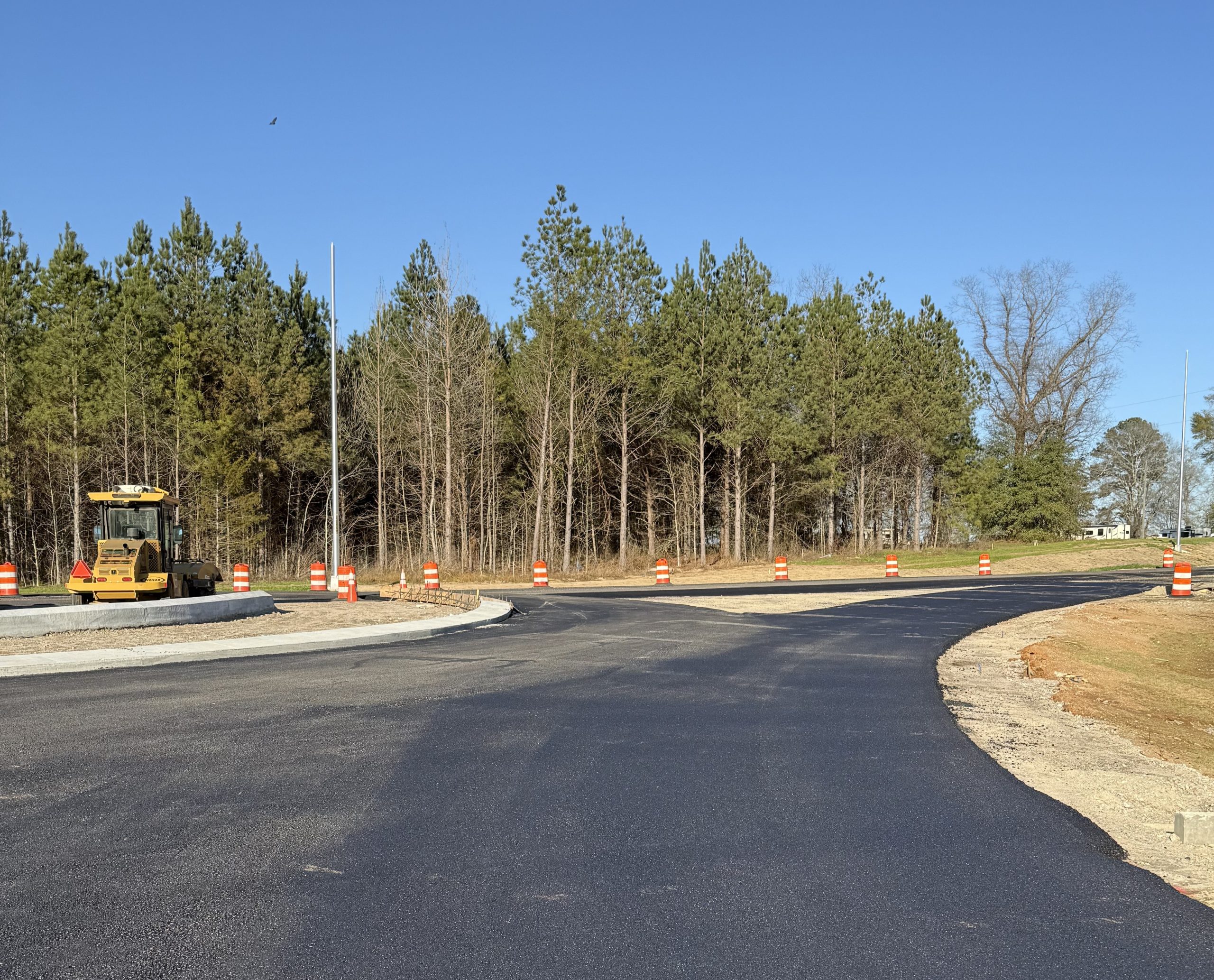 Freshly paved asphalt at the US-80 and Marler Road roundabout construction site, featuring orange traffic barrels and a yellow steamroller against a treeline.