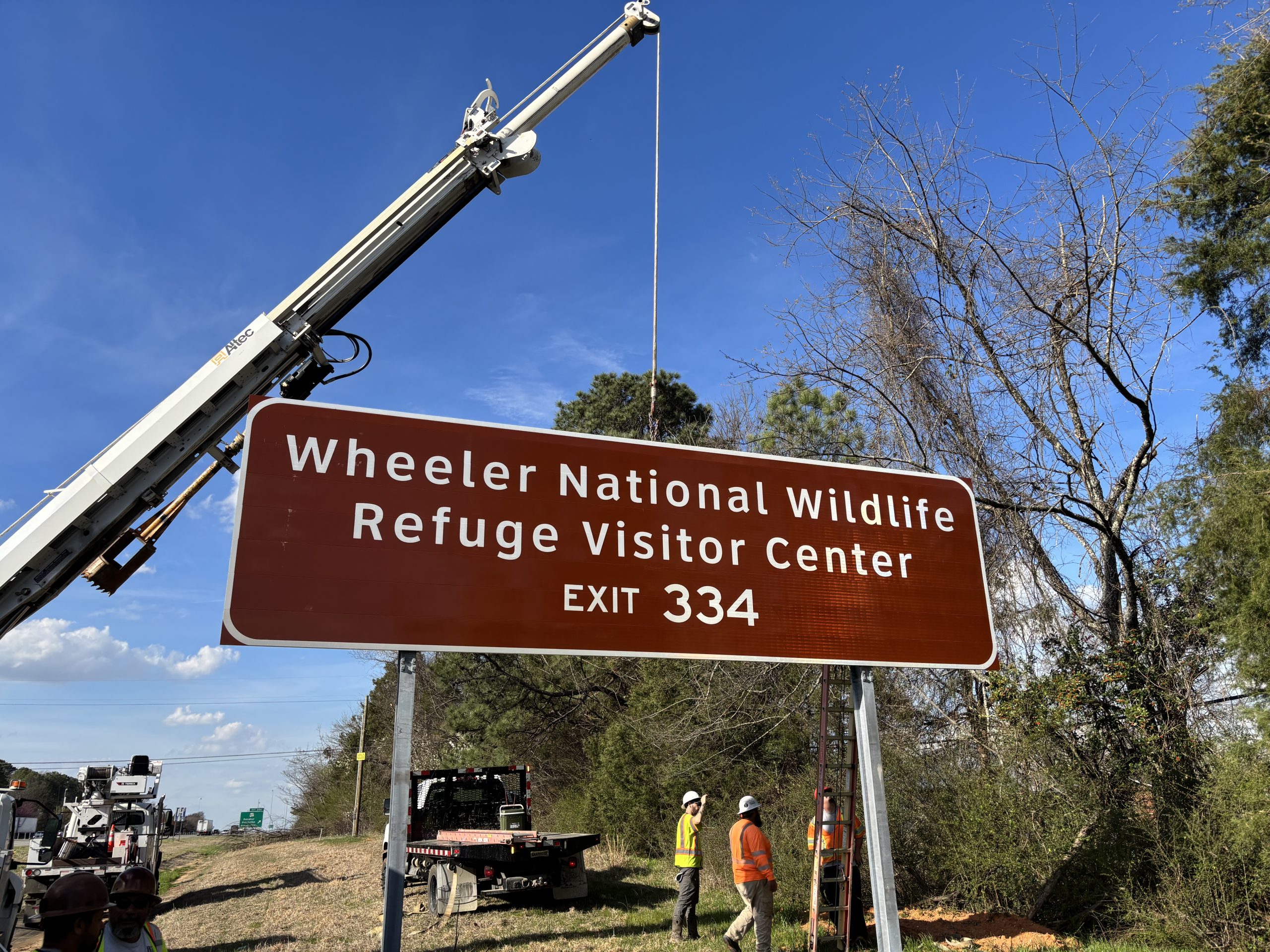A brown sign for Wheeler Wildlife Refuge Visitor Center Exit 334, suspended from a crane, is fastened to sign poles.