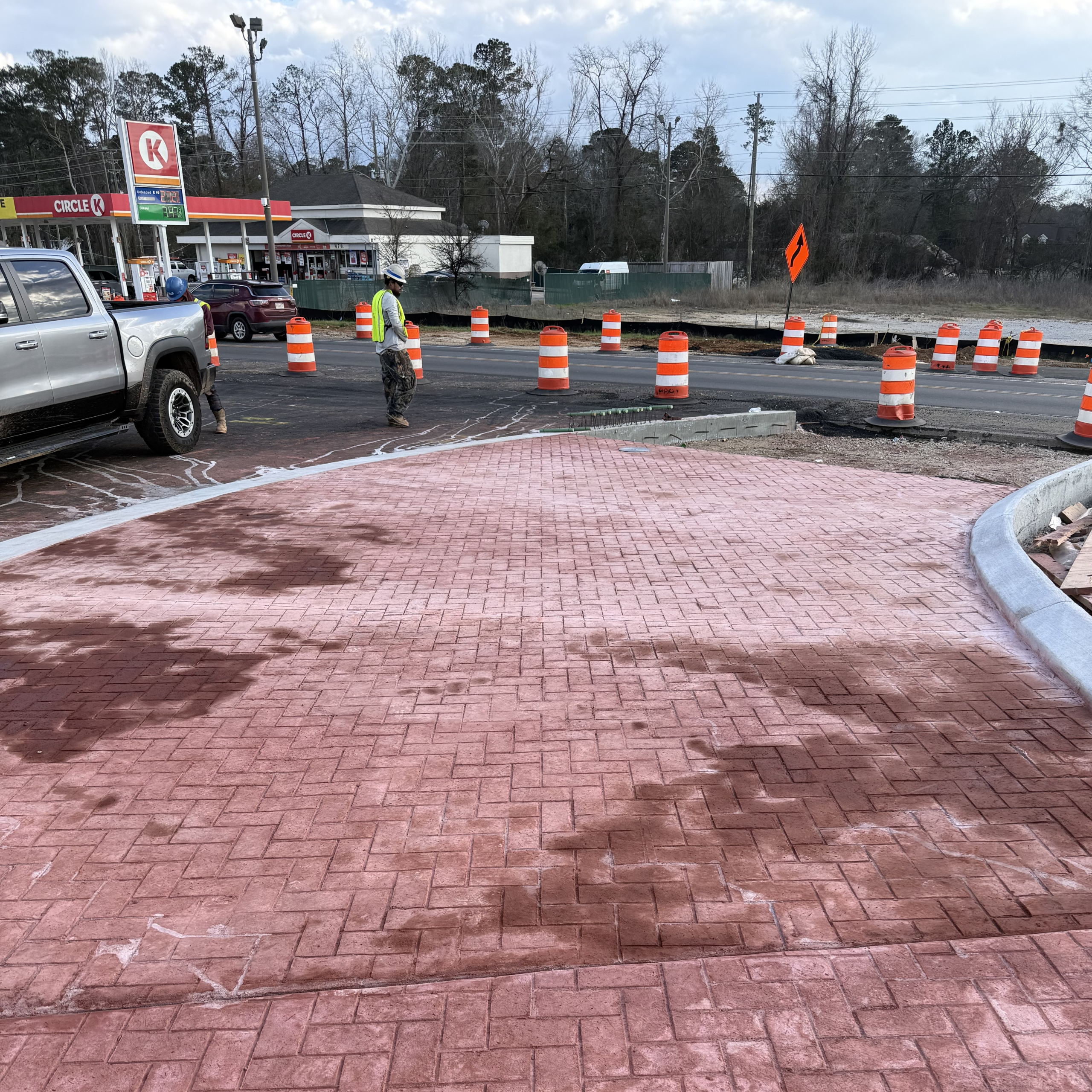 A construction crew worker in a high-visability vest stands near a new red brick-patterned island. In the background, orange traffic barrels line a roadway near a gas station at US-80 and SR-126.