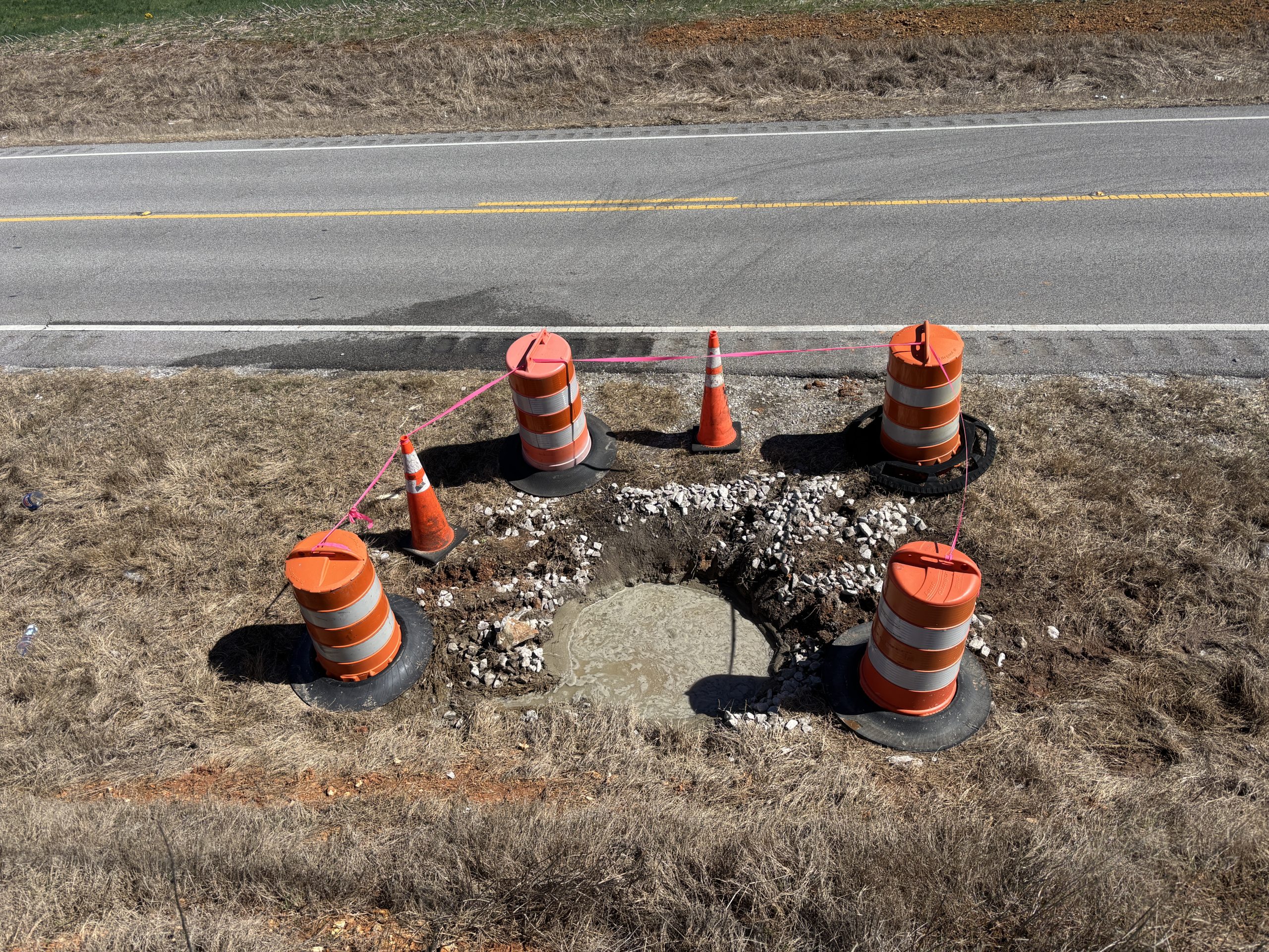 A circular depression filled with concrete and surrounded by drums and cones near a two-lane highway.