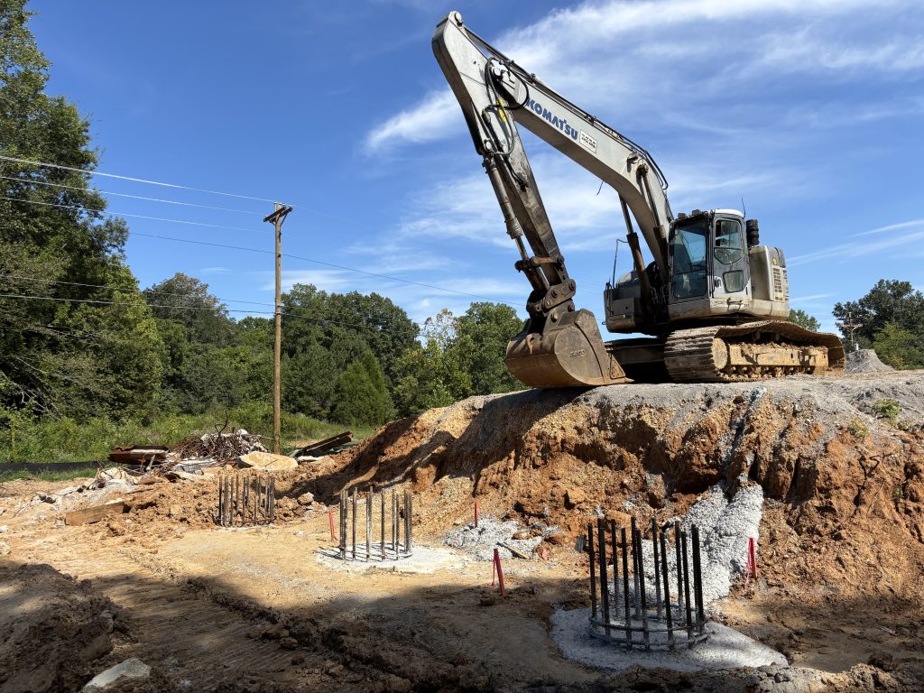 An excavator perched on a mound of earth over three circles of concrete with rebar sticking up.
