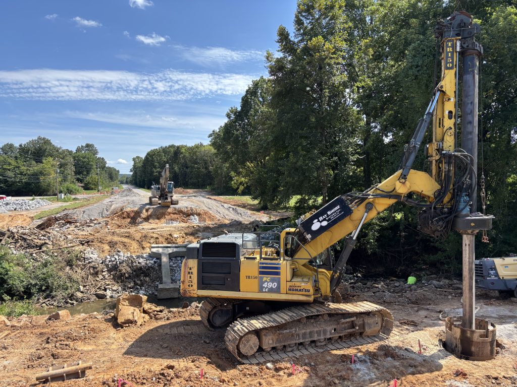 An auger drilling into the earth. An excavator can be seen on the other side of the creek.