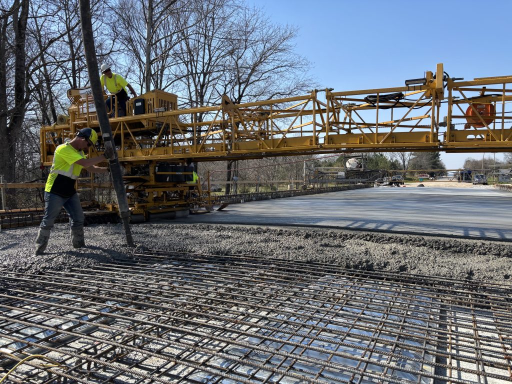 A worker with a boom and concrete pouring out while a machine  levels the concrete behind him