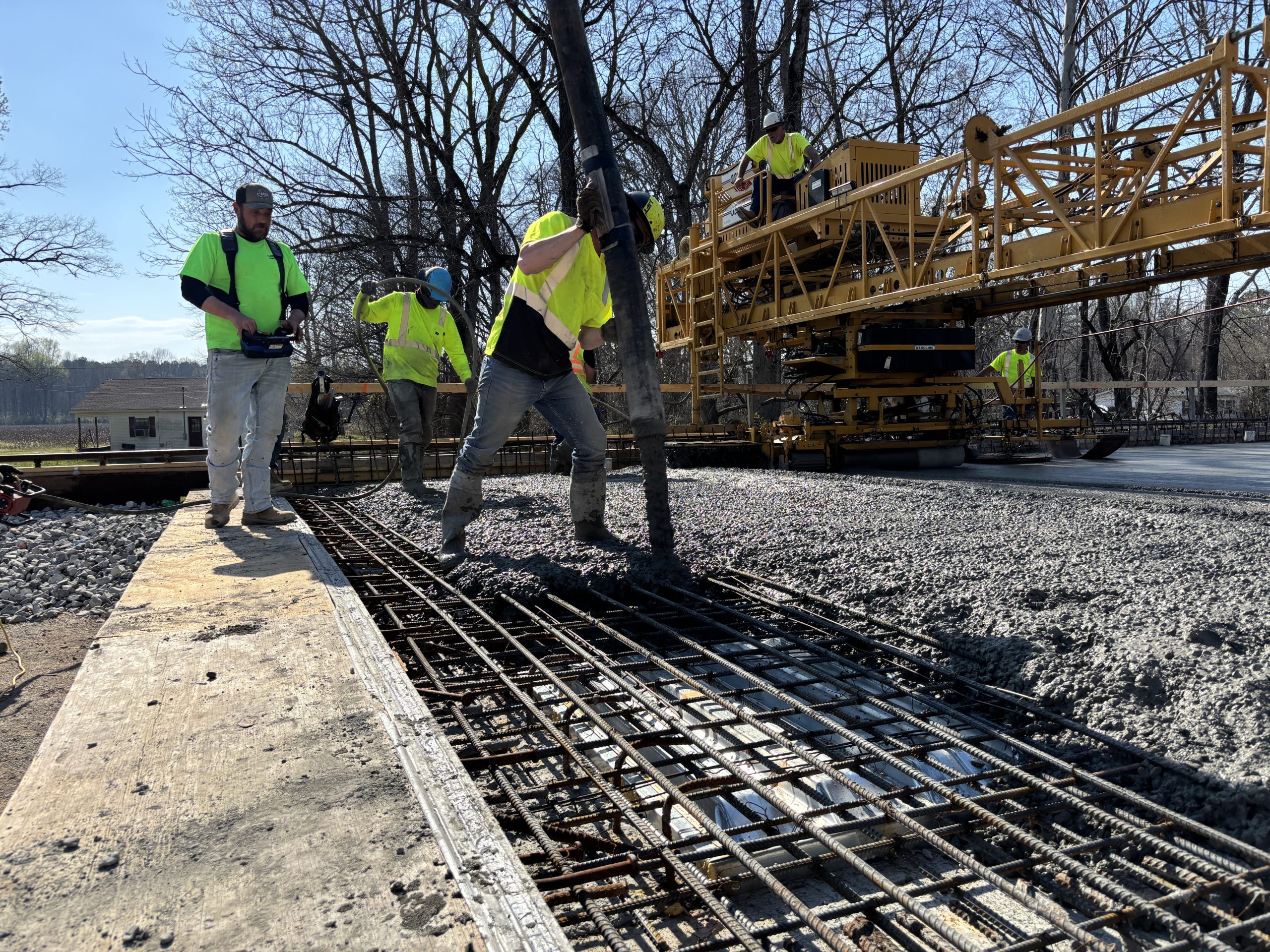 Three workers on a grid of steel rebar surrounded by wet concrete. One directs the boom by hand while another uses remote controls to operate the boom truck. A gantry is visible in the background.