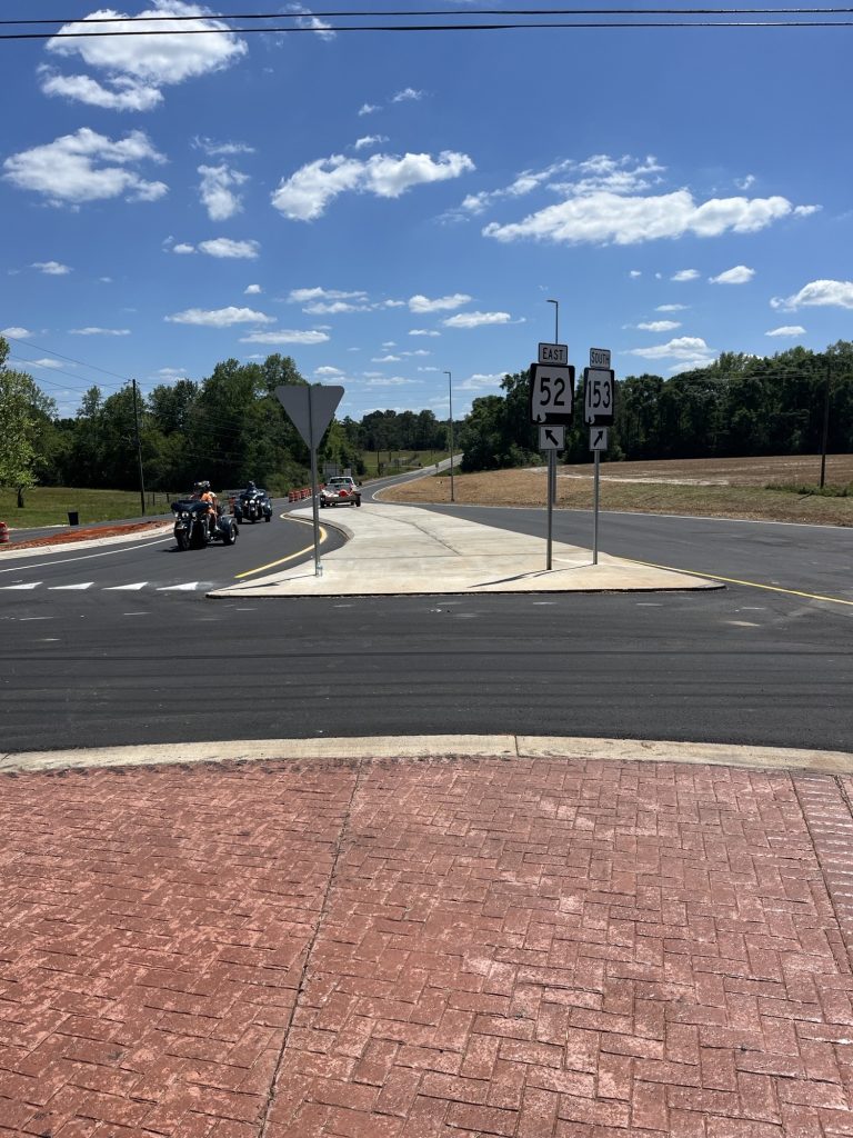 An intersection featuring a new roundabout under a bright blue sky with scattered clouds. In the foreground, red-brick patterned pavement transitions into fresh black asphalt. Traffic signs point toward East SR-52 and South SR-153. Motorcyclists and a white pickup truck are visible on the curving roadway, which is lined with green trees and open fields.
