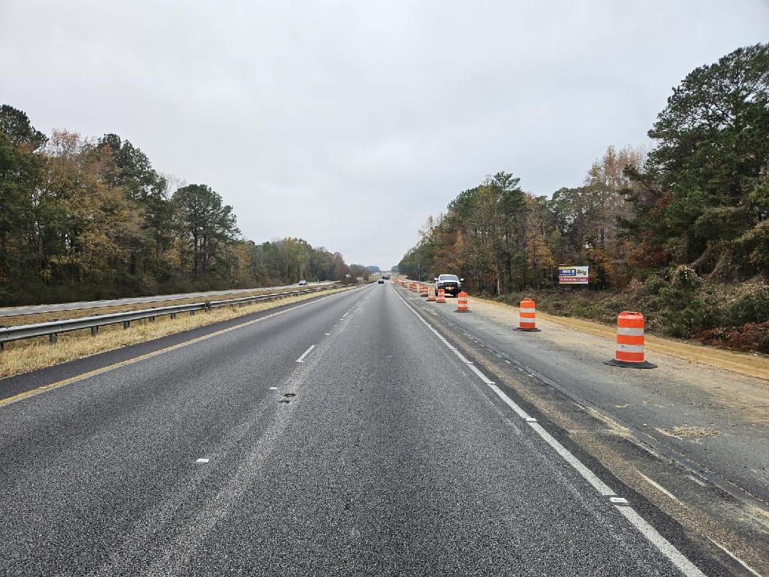 A view of a construction zone on I-85 South near Miles Creek Bridge, featuring orange and white traffic barrels lining the shoulder of the highway. The road stretches toward a distant horizon under an overcast sky with dense autumn foliage on either side.