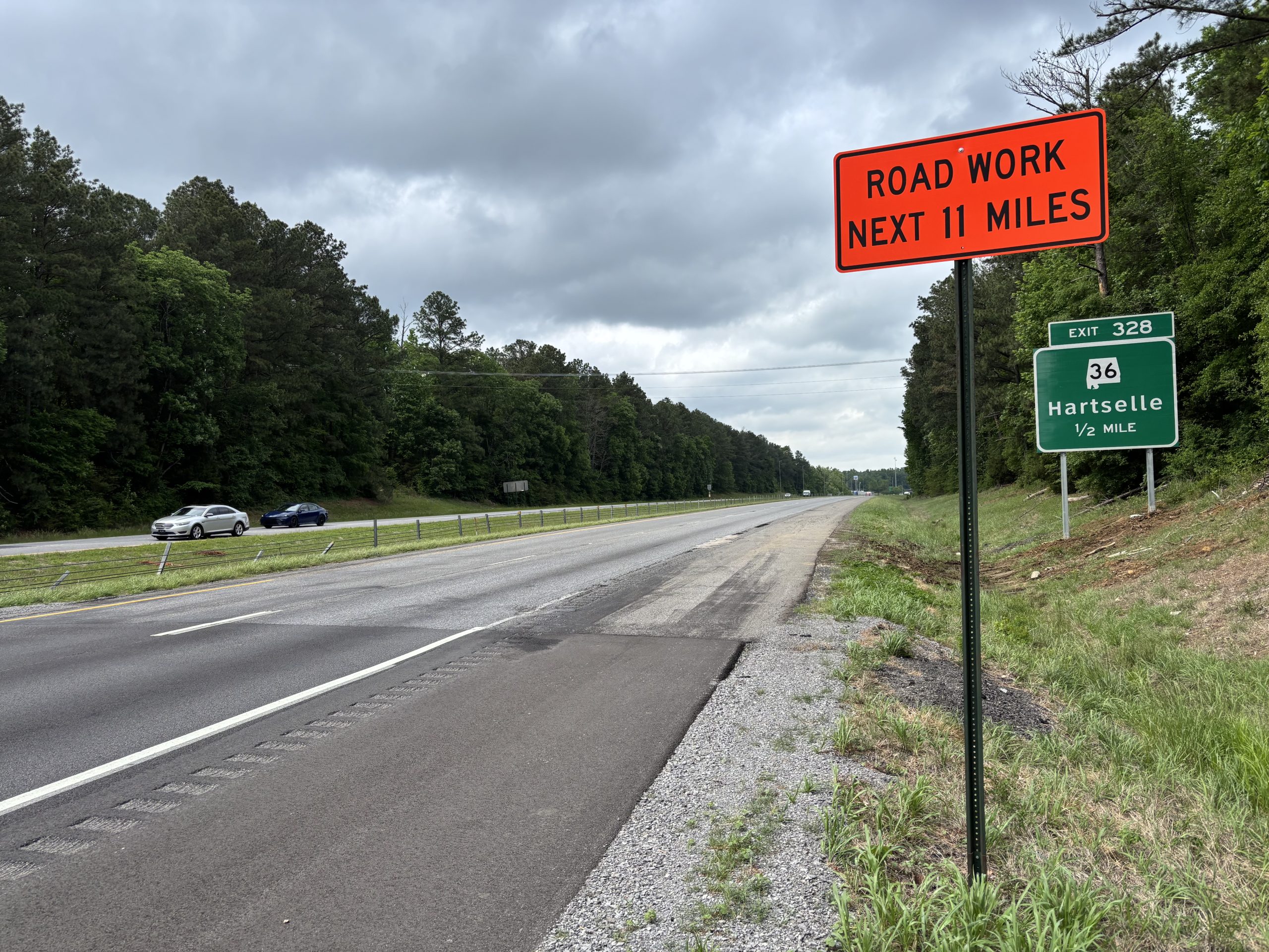 A joint at the end of a recently-resurfaced segment of interstate. The poorer condition of the old pavement past the joint is apparent. An orange sign posted on the shoulder reads: RTOAD WORK NEXT 11 MILES. A green Exit sign identifies the upcoming Exit as Exit 328 at SR-36 in Hartselle,