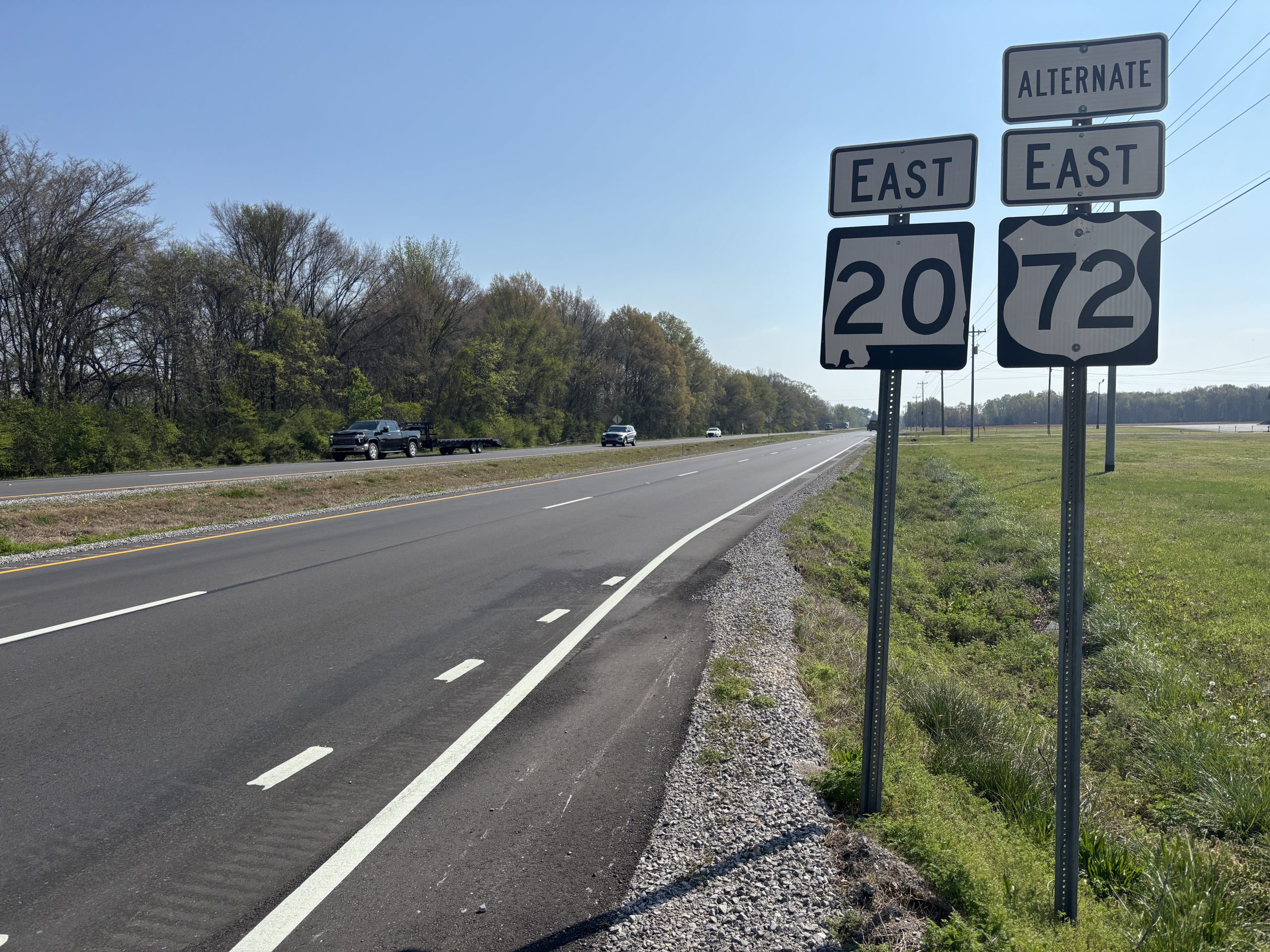 A recently paved road with a pair of signs posted on the shoulder indicating it is eastbound State Route 20 and also US Alternate 72.