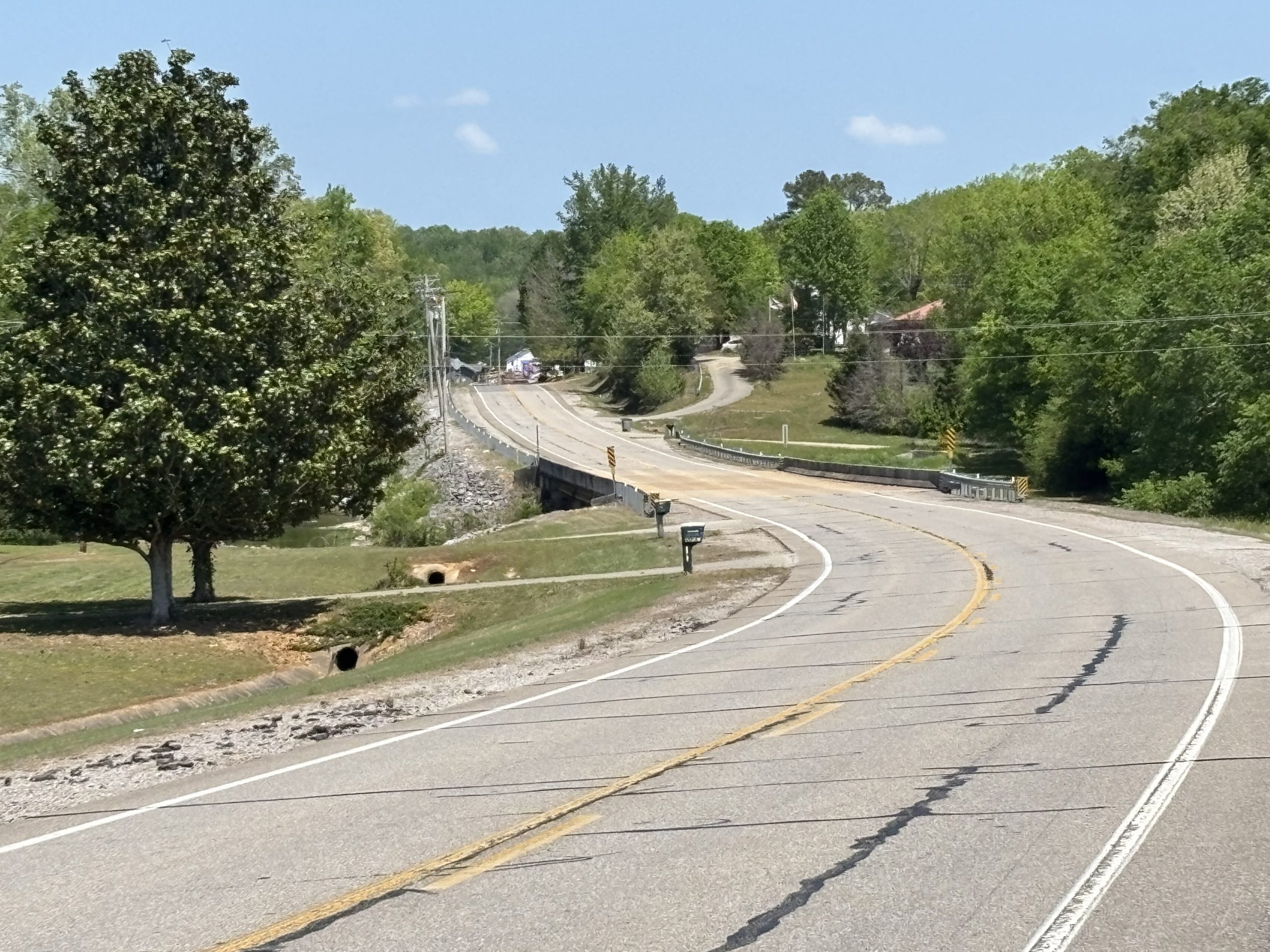 A two-lane road winds across a creek. Signs of previous repairs and crack sealing are visible in aging pavement surface.