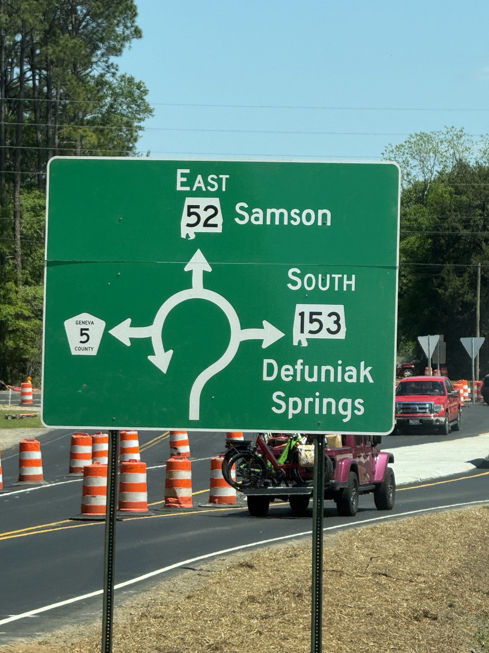 A large green highway directional sign for the SR-52 roundabout. The sign features a white diagram of the roundabout with arrows pointing toward East 52 Samson, South 153 Defuniak Springs, and Geneva County 5. In the background, orange construction barrels line a freshly paved road where a pink Jeep and a red pickup truck are driving.