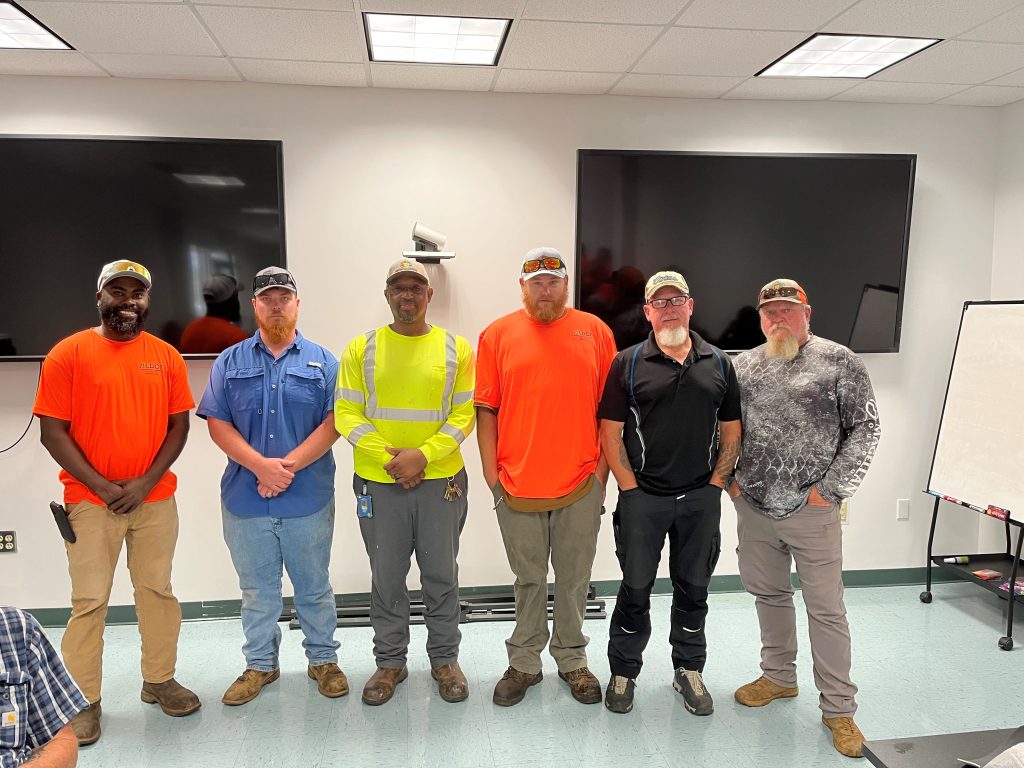 Six crew members in casual and high-visibility workwear stand in a row for a group photo inside an office or training room.