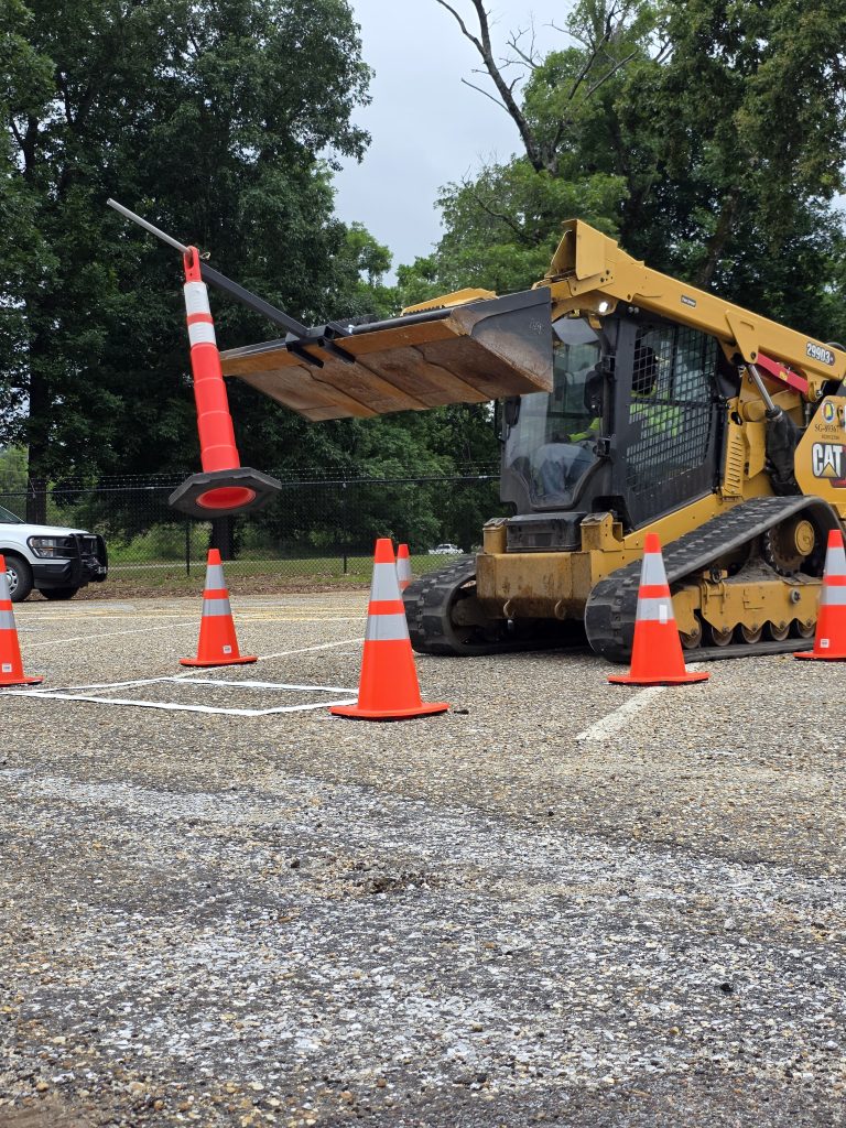 A skid steer operator uses a specialized attachment on the bucket to lift and move a traffic delineator post with extreme precision during a skills competition.