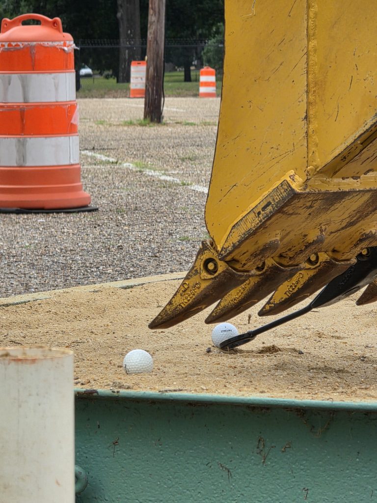 A backhoe bucket demonstrates precision by using a small spoon attachment to carefully place a golf ball into a sand pit.