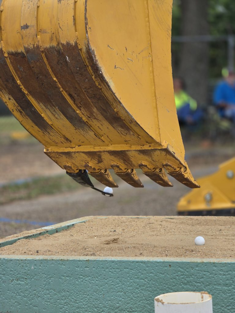 An excavator bucket carefully balances a golf ball on a small spoon attached to one of its teeth, demonstrating precision during a training exercise.