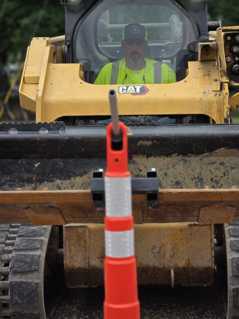 An equipment operator maneuvers a skid steer to align a metal rod with the top of a traffic delineator post, showcasing technical skill and accuracy.