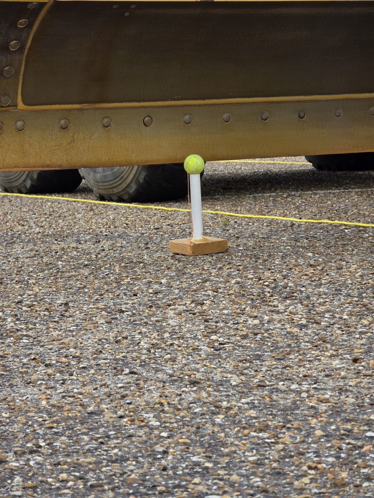 A tennis ball remains balanced on a stand as the massive blade of a motor grader passes just inches above it, demonstrating the incredible control and precision of the operator.