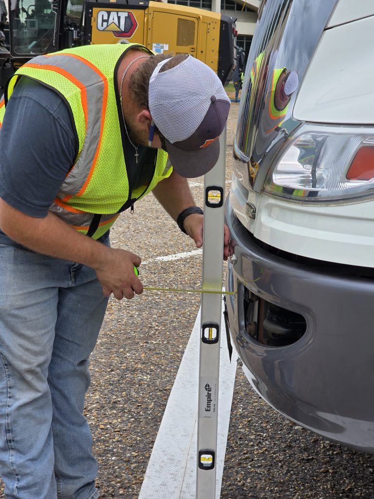 A crew member in a safety vest uses a level and tape measure to perform a precise measurement on the front bumper of a white truck.