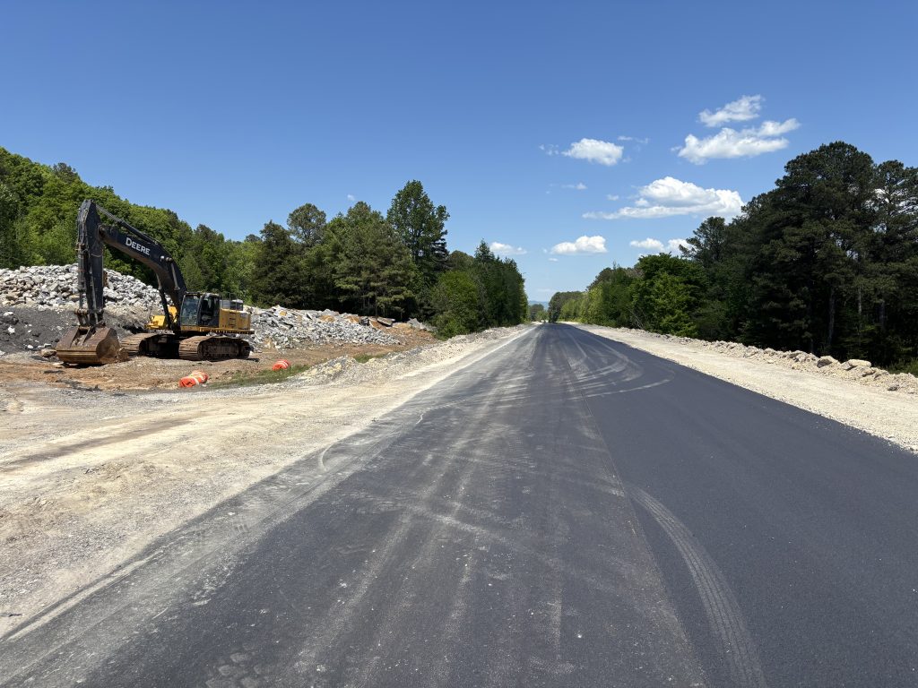 A roadway with an excavator parked next to it. In the median is a large pile of broken concrete slabs.