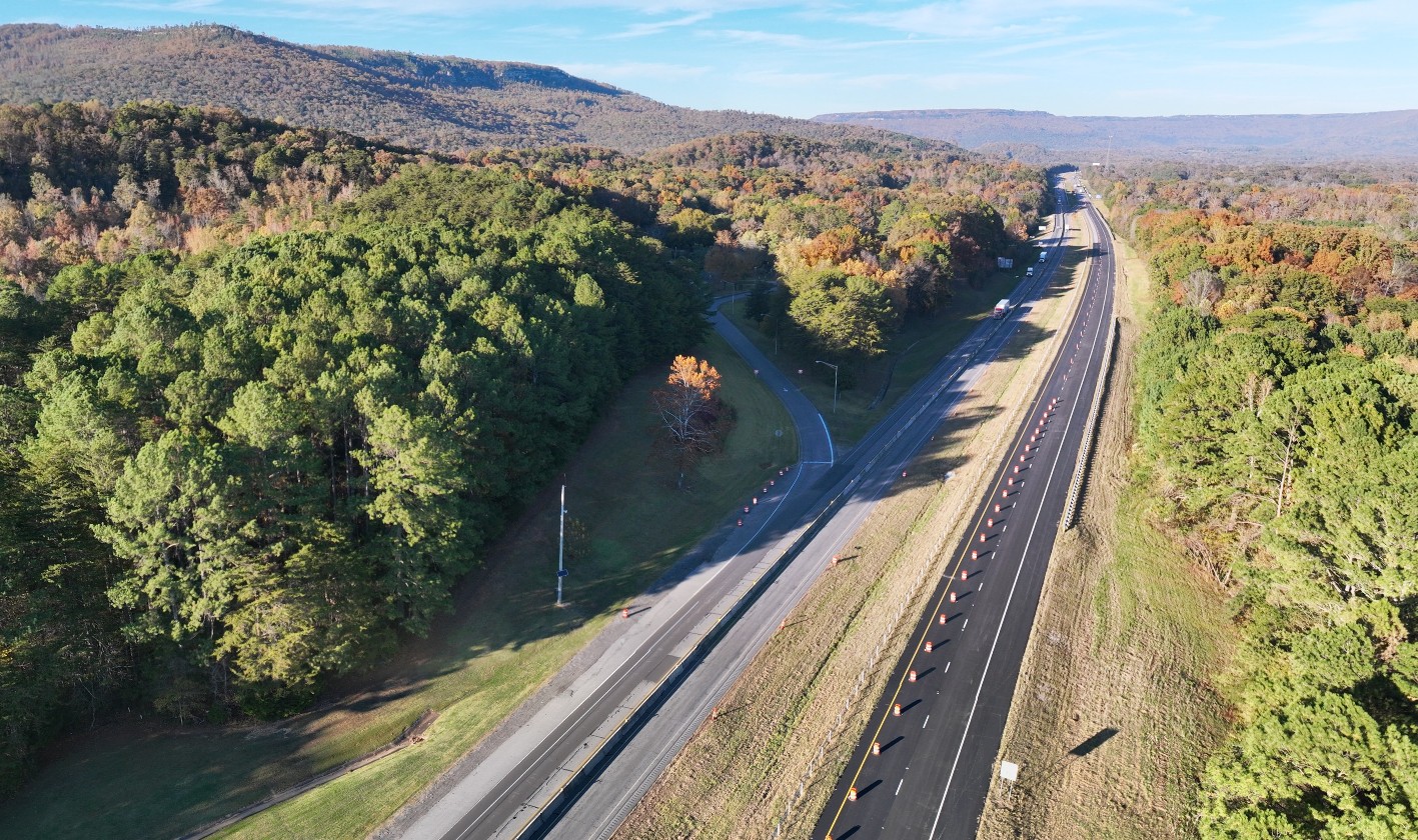 An aerial view looking north at an interstate highway. The northbound road to the right has recently been reconstructed. The southbound road to the left is carrying two-way traffic diverted from the northbound road. An on-ramp merges to the southbound roadway. from a welcome center that is covered by the trees.