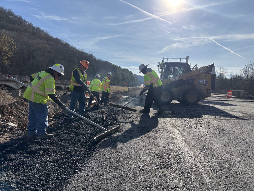 Workers level asphalt placed by a skid steer at the the edge of a road at the end of an overpass.