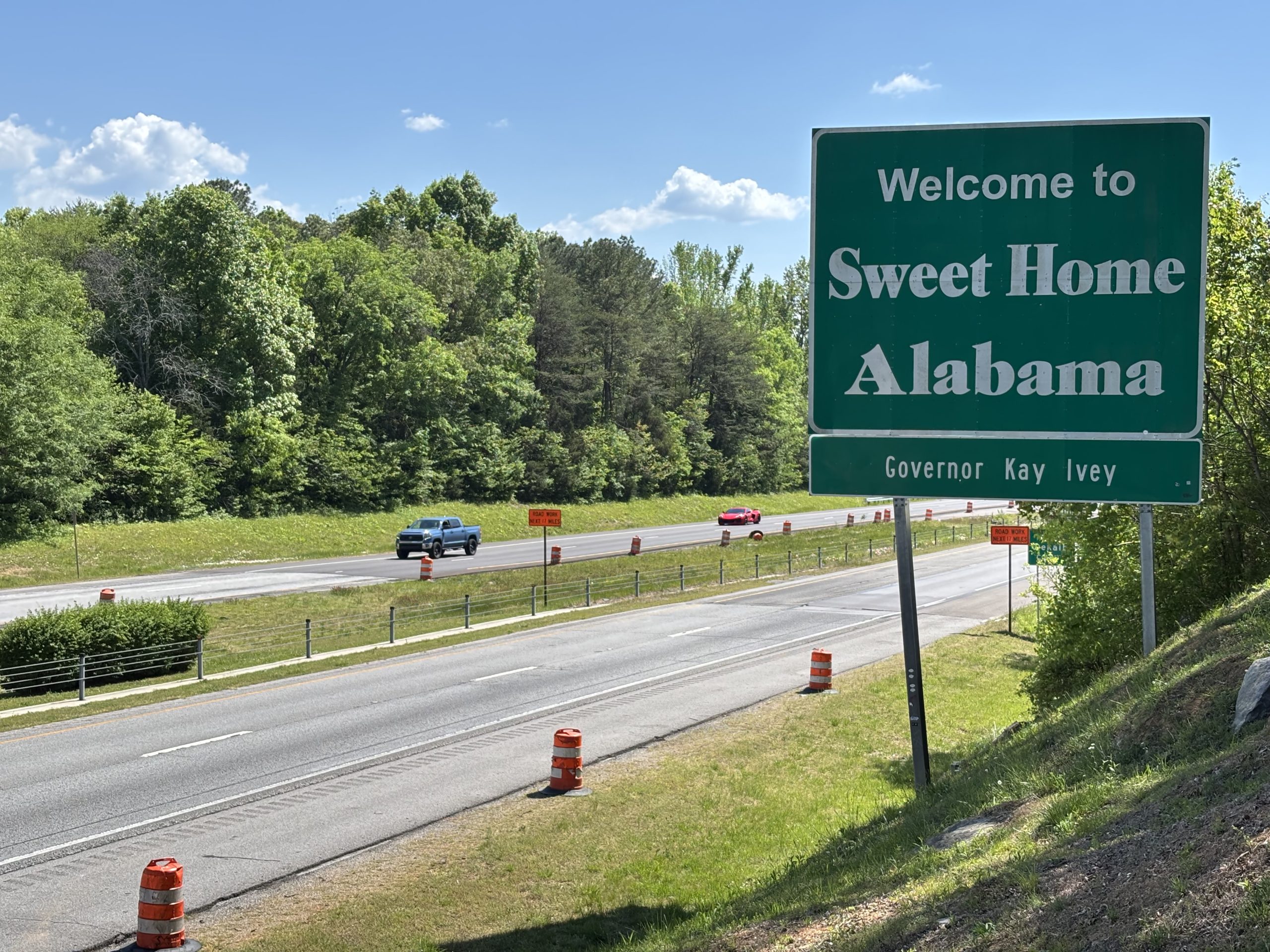 Traffic drives past a large green "Welcome to Sweet Home Alabama" sign. Orange and white drums line the roadway.