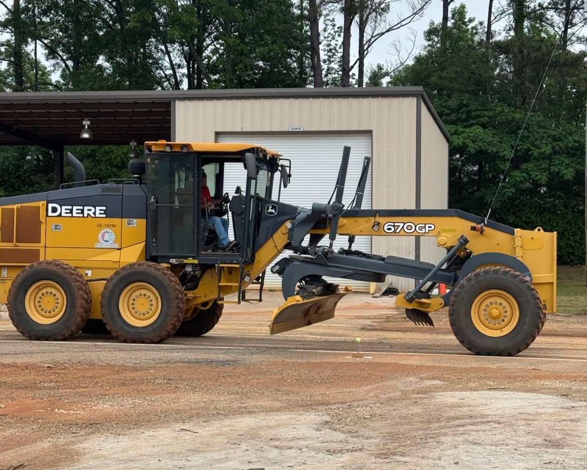 A John Deere motor grader operator navigates a precision course, carefully maneuvering the large blade over a small target on the ground.