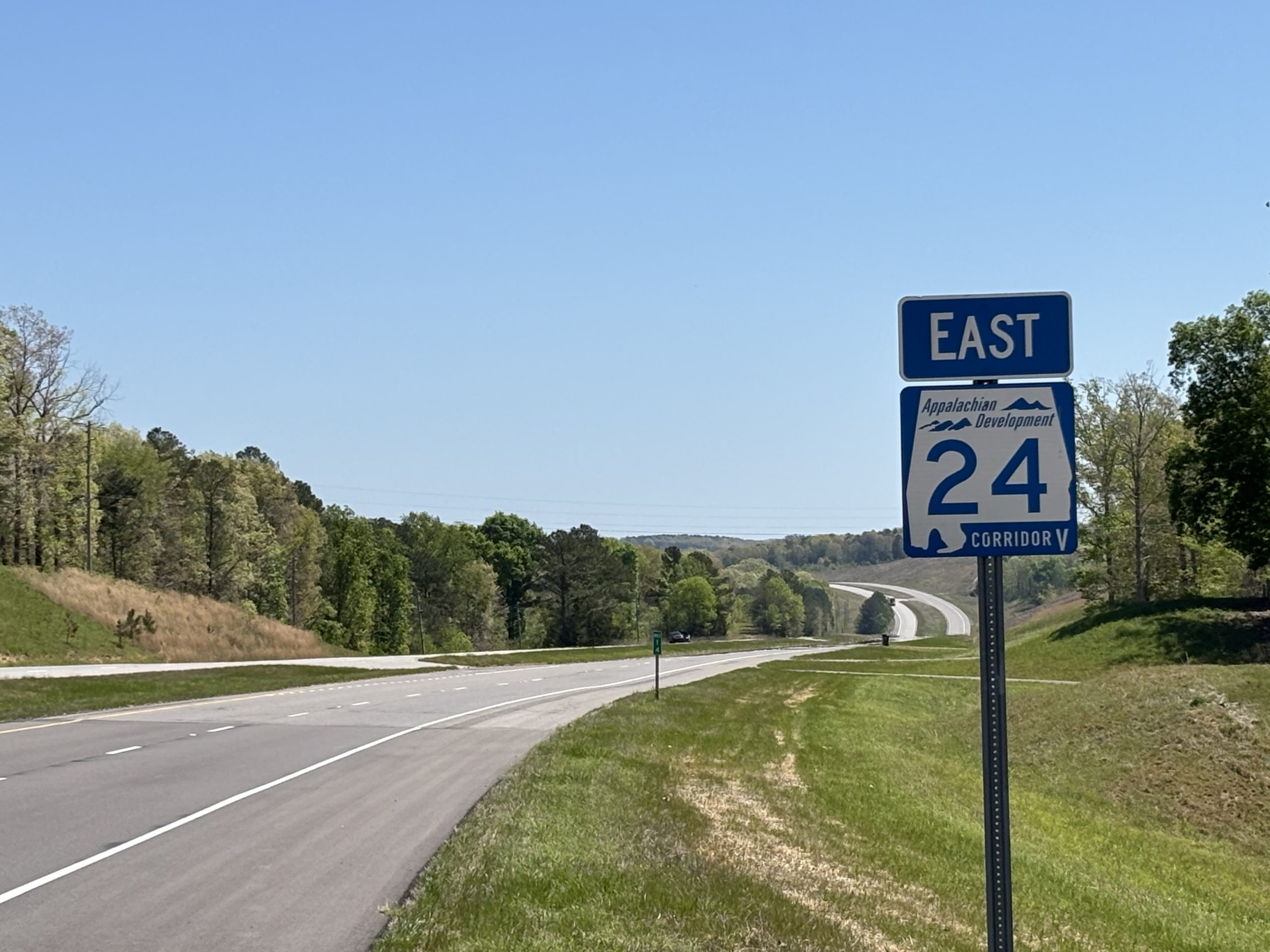 A blue and white state route shield marks SR-24 as an Appalachian Development Corridor. The divided four-lane highway winds over hills. A patched area of pavement is visible.