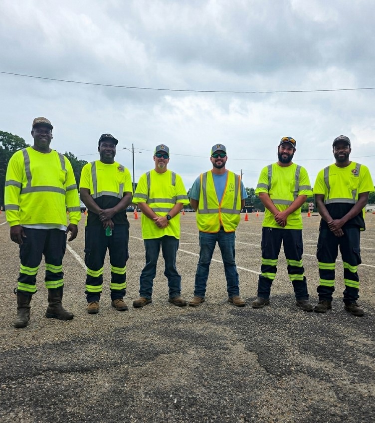 Six crew members in high-visibility safety workwear stand in a row across an asphalt lot during an outdoor training event.