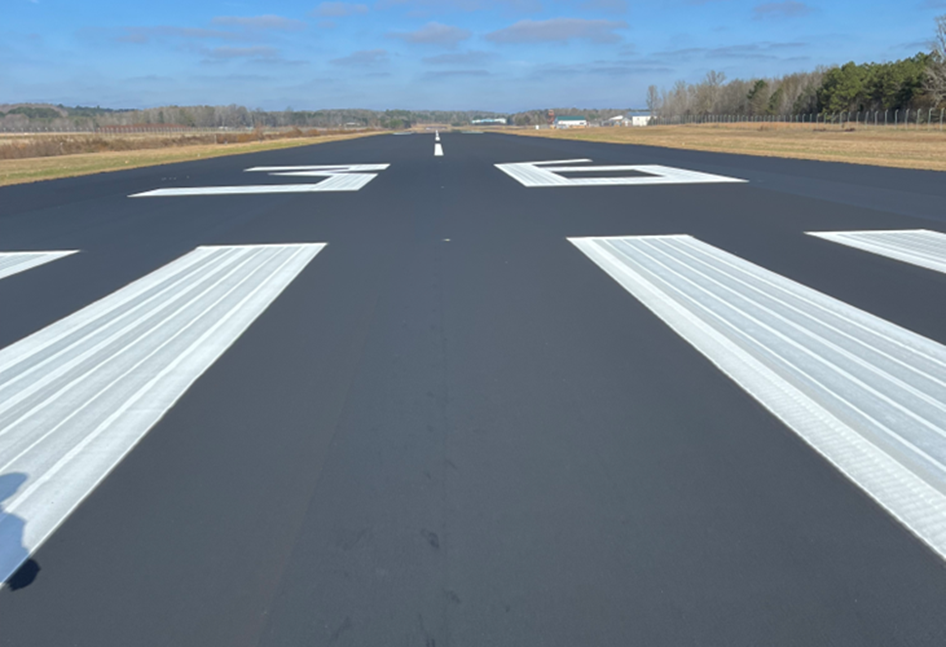 Freshly resurfaced airport runway with newly painted white threshold markings and centerline stripe extending into the distance. The smooth black pavement is bordered by grass and trees under a partly cloudy sky.