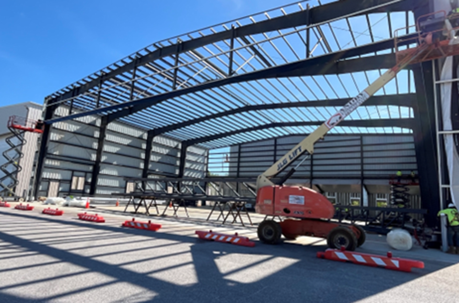 Steel framework of a large aircraft hangar under construction at an airport. Workers use aerial lifts inside the structure as roof supports and wall panels are installed. Construction materials and safety barriers are visible on the concrete apron beneath a clear blue sky.