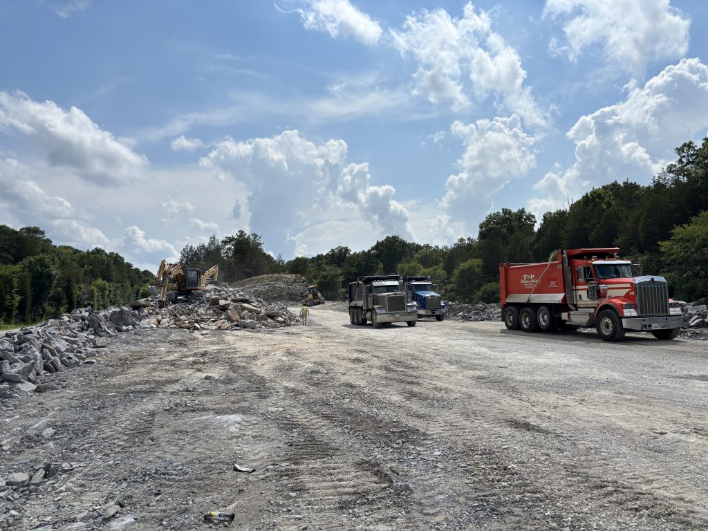 Dump trucks haul rock away from a site where excavators are working.