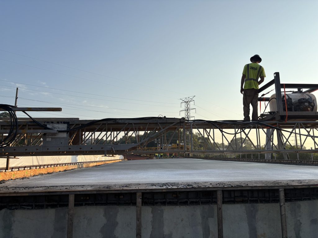 A worker on a gantry silhouetted by the morning sun above a freshly-poured concrete bridge deck