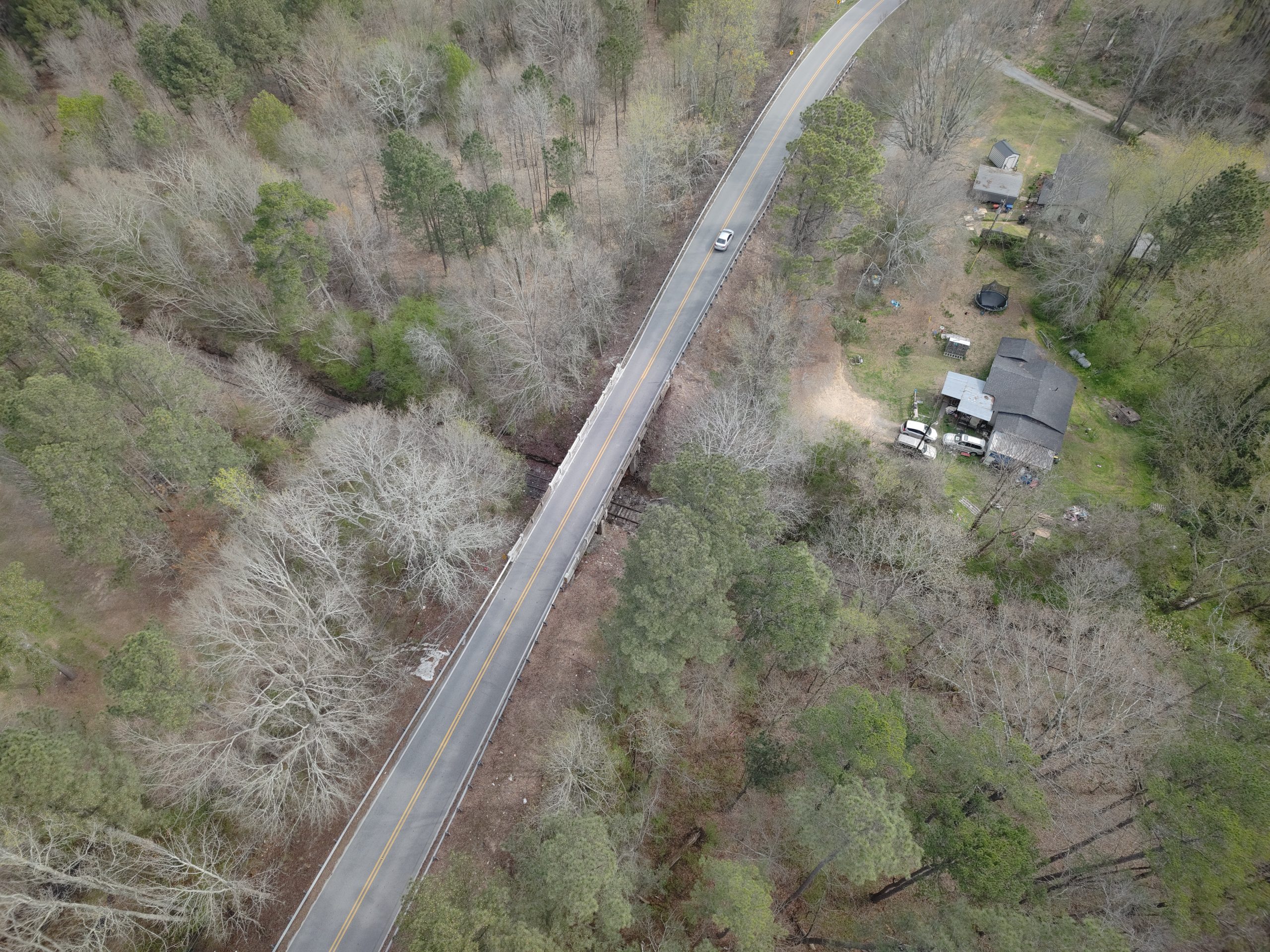An aerial view of a road with a bridge over a railroad tracks, surrounded by trees