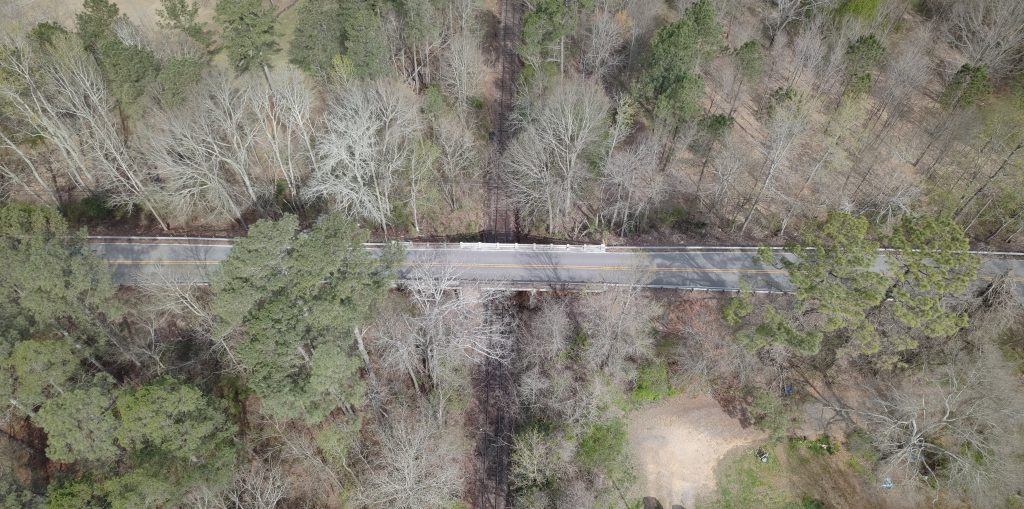 An overhead view of a road with a bridge over a railroad tracks, surrounded bytrees